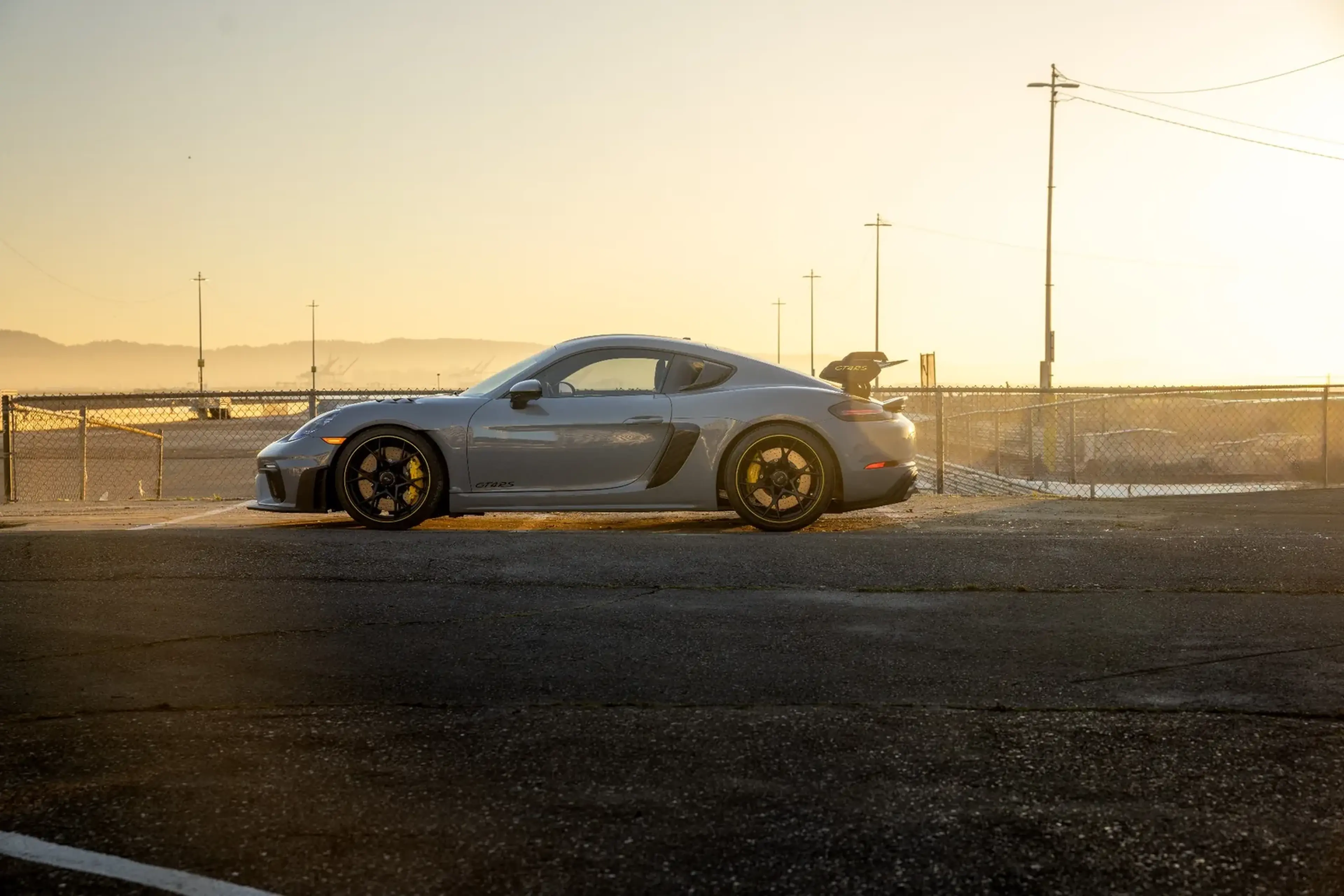 A silver sports car parked on a sunlit road near a fence, with a clear horizon and streetlights in the background.