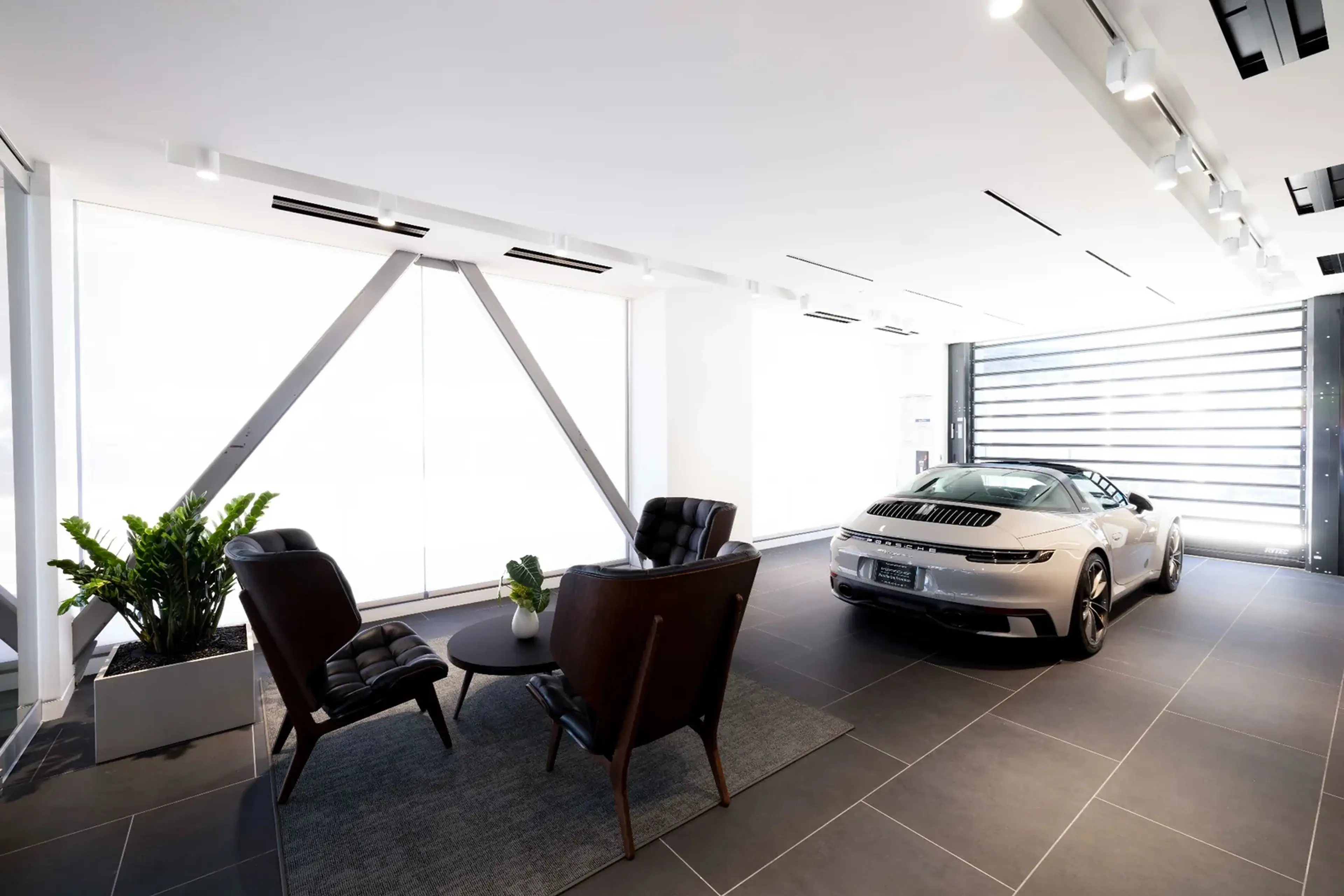 Luxury car in a stylish showroom next to a seating area with two leather chairs, a round table, and potted plants.