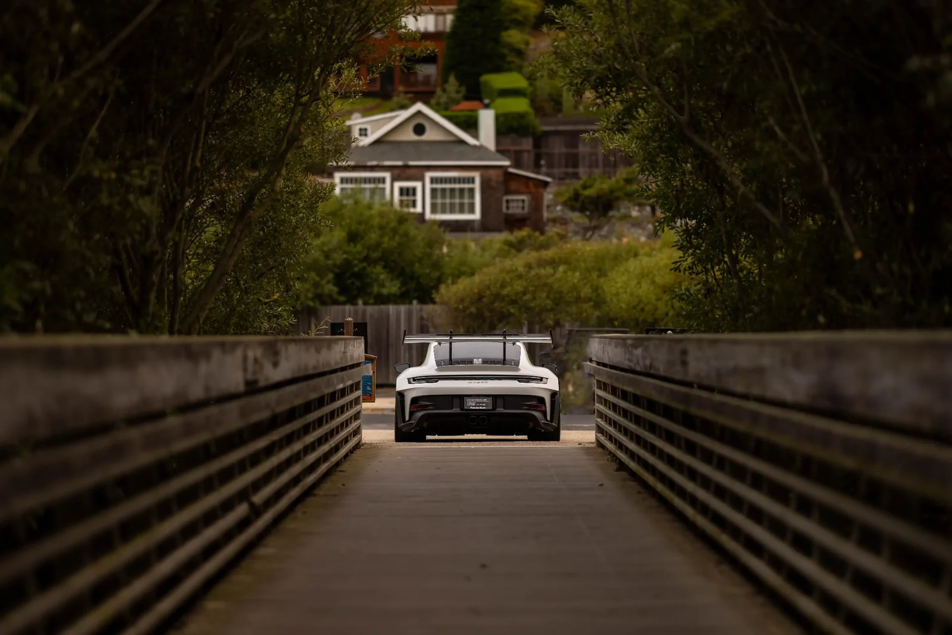 A white sports car with a large rear wing is parked on a bridge leading to a house surrounded by greenery.