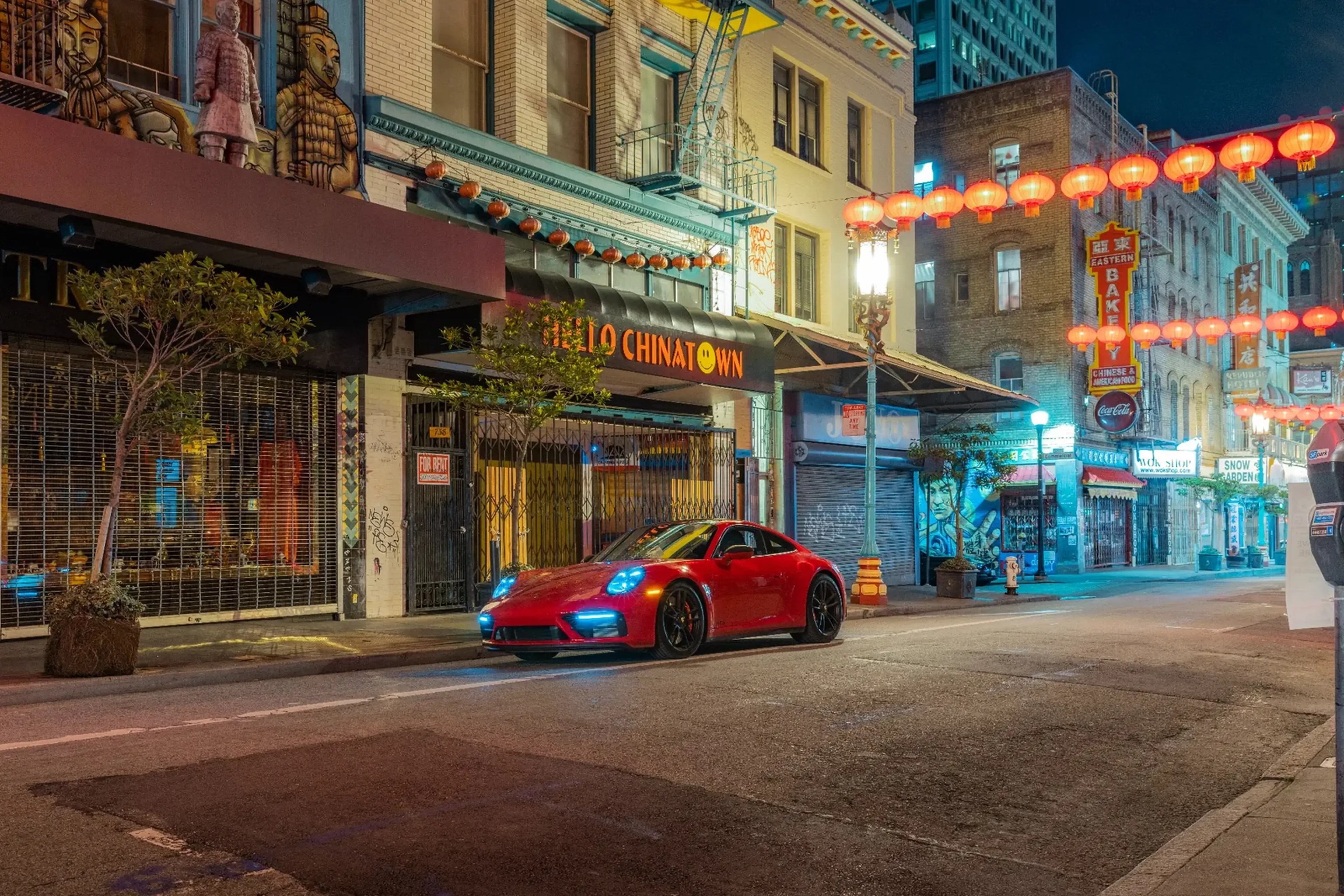 A red sports car is parked on a quiet street in Chinatown, adorned with red lanterns and colorful storefronts at night.