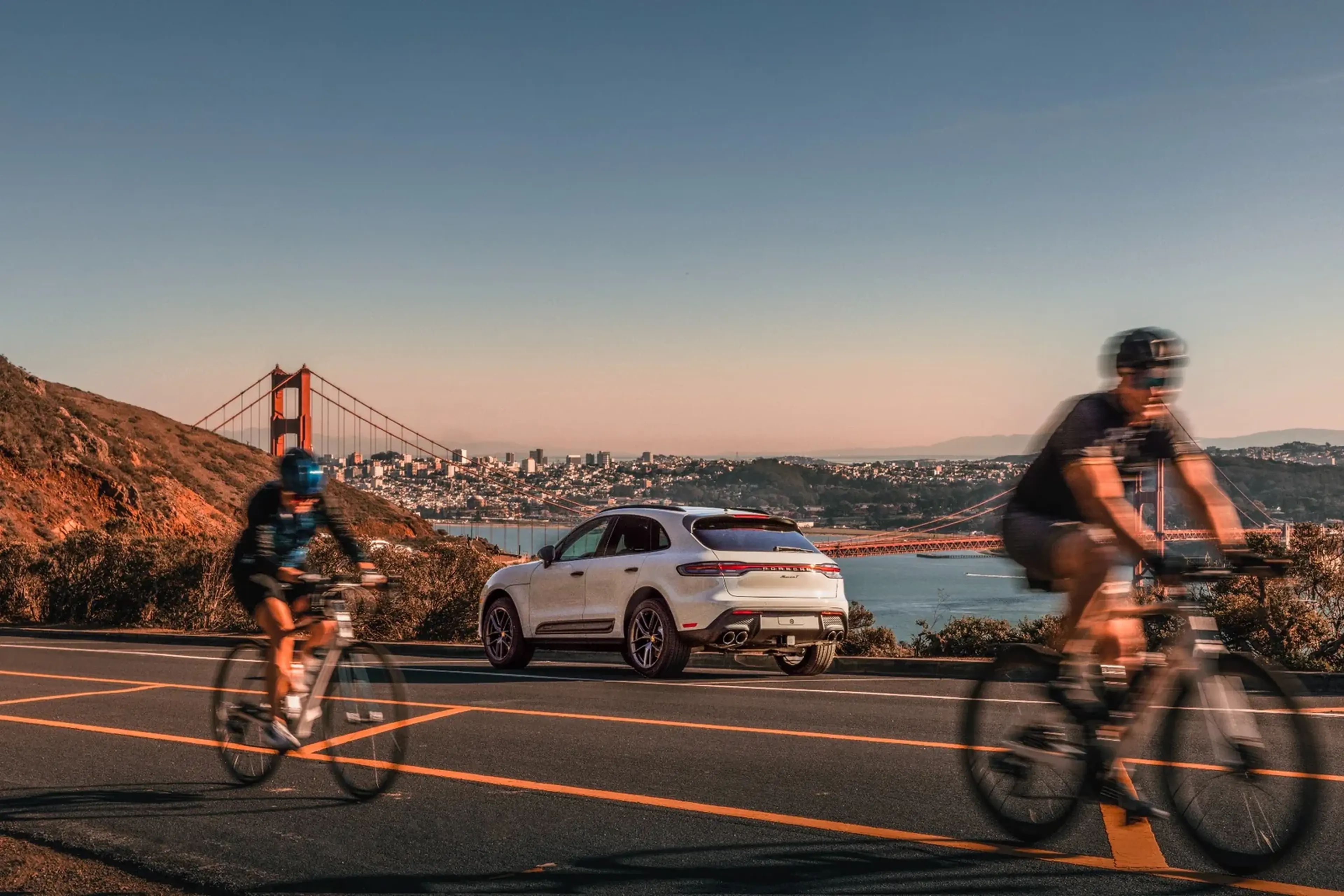 Two cyclists ride past a parked white SUV with the Golden Gate Bridge and San Francisco cityscape in the background under a clear sky.