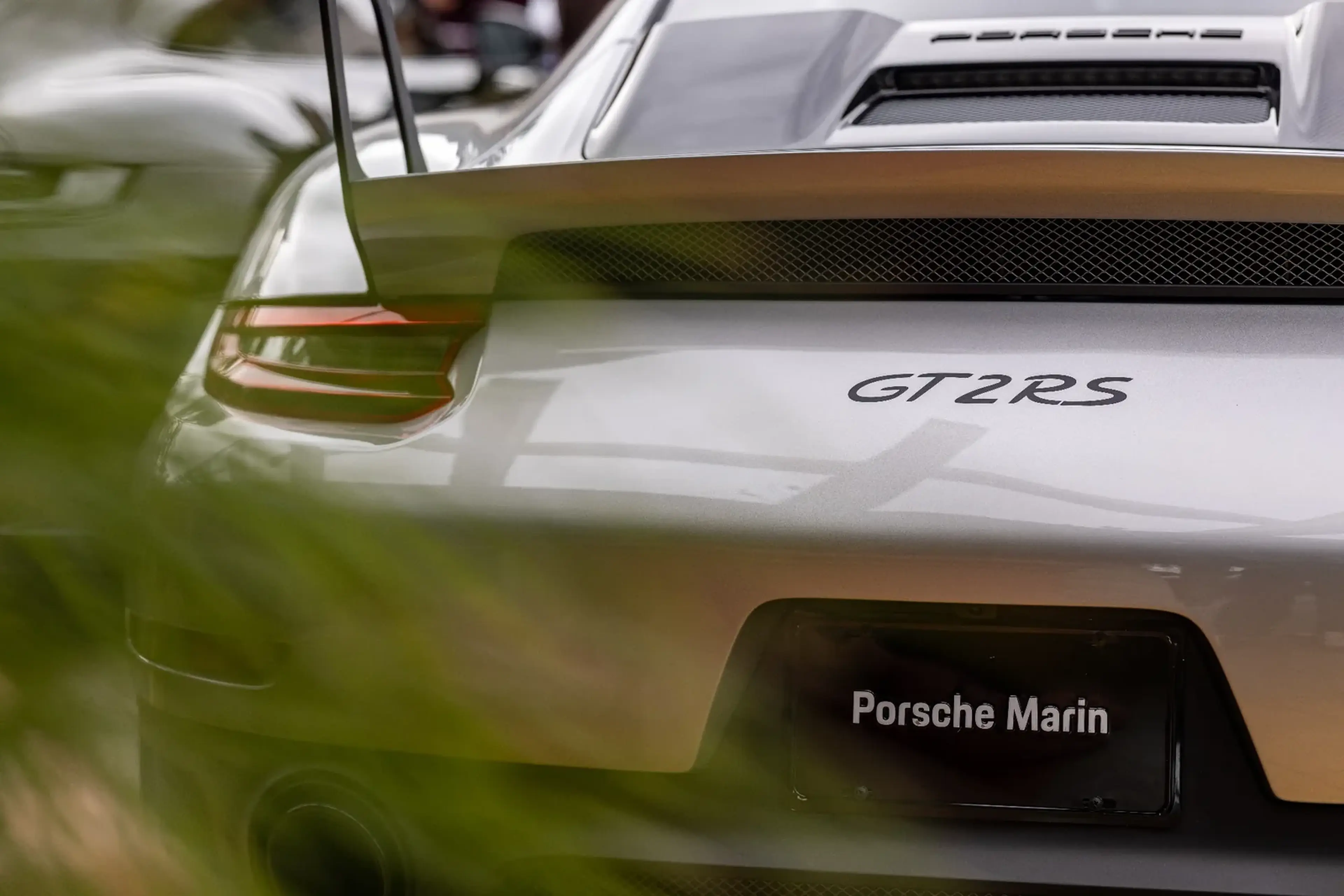 Rear view of a white Porsche GT2 RS with a spoiler, partially obscured by green foliage.