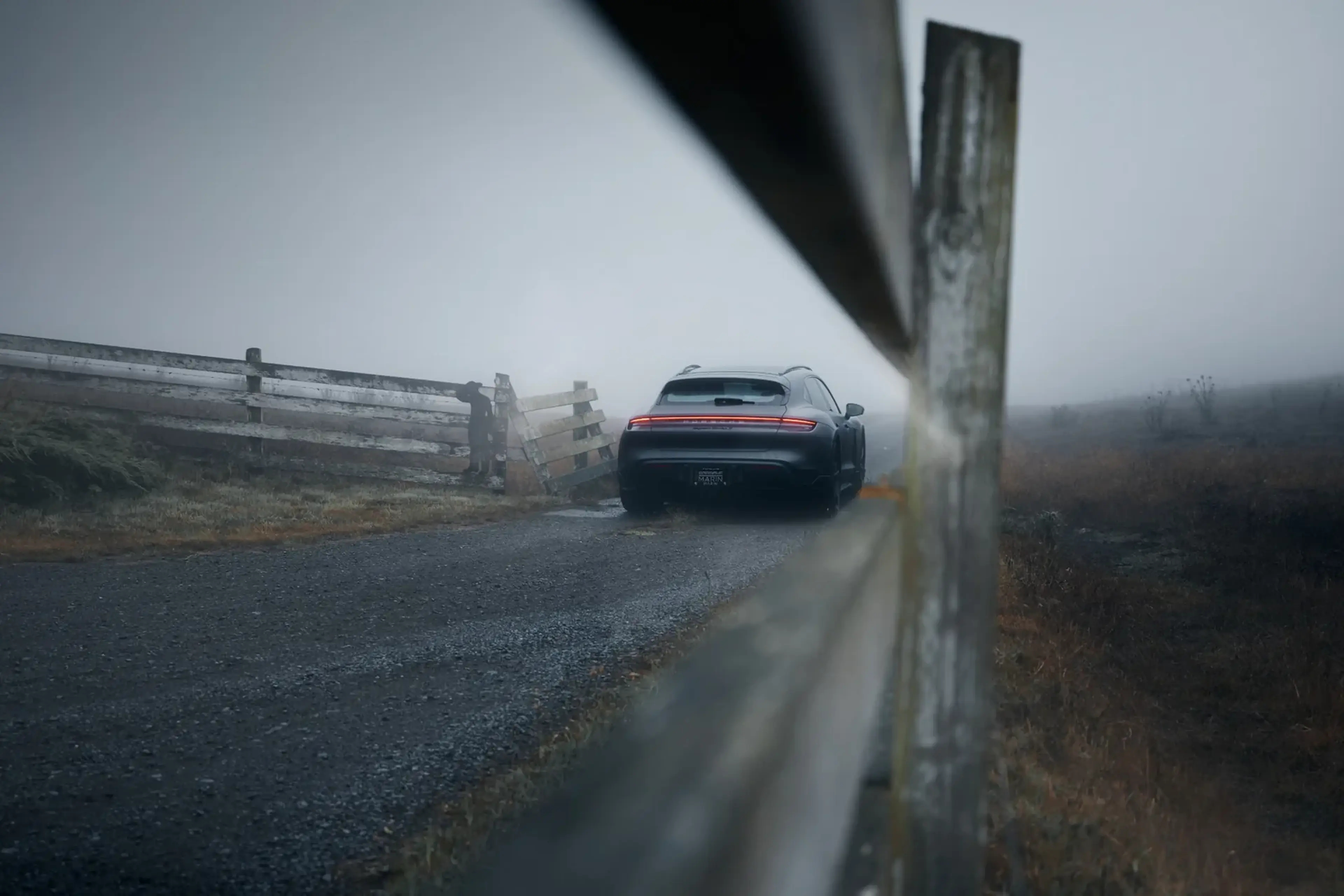 A car with illuminated taillights is parked on a foggy rural road between parallel wooden fences.