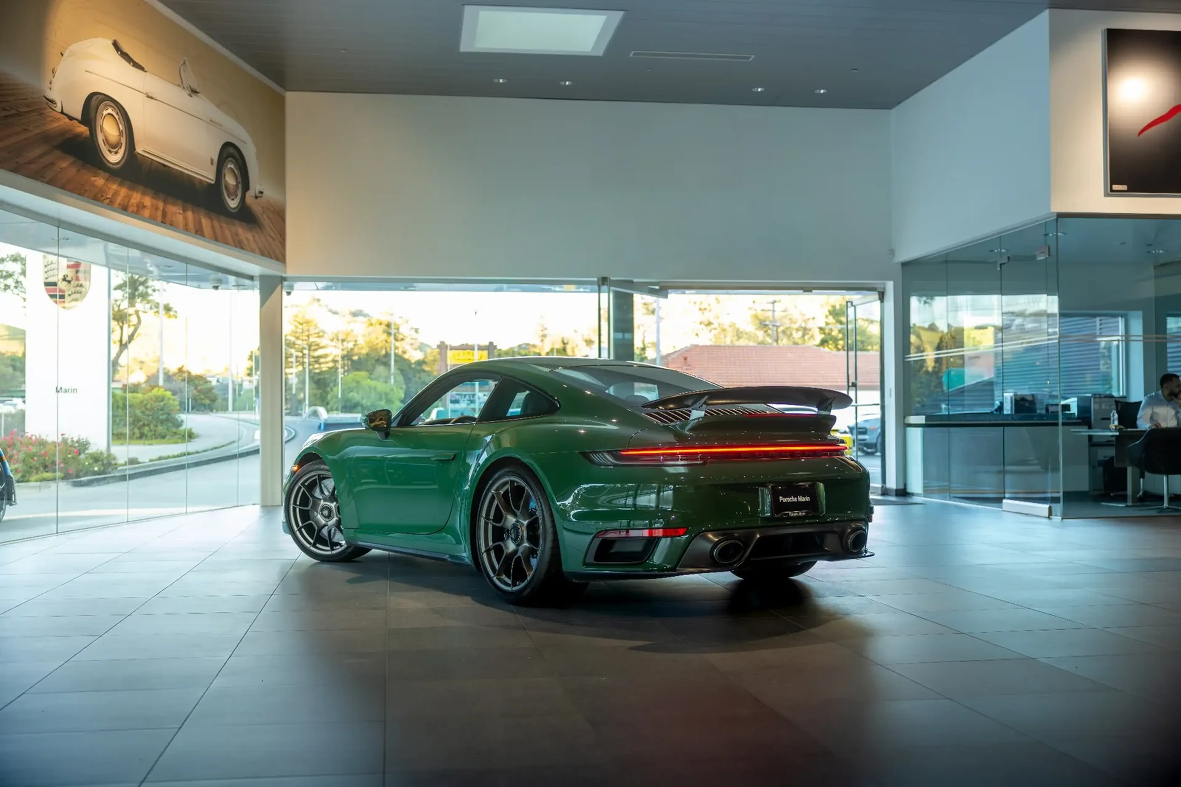 Green sports car displayed in a showroom with large windows, featuring a sleek rear design and black rims.