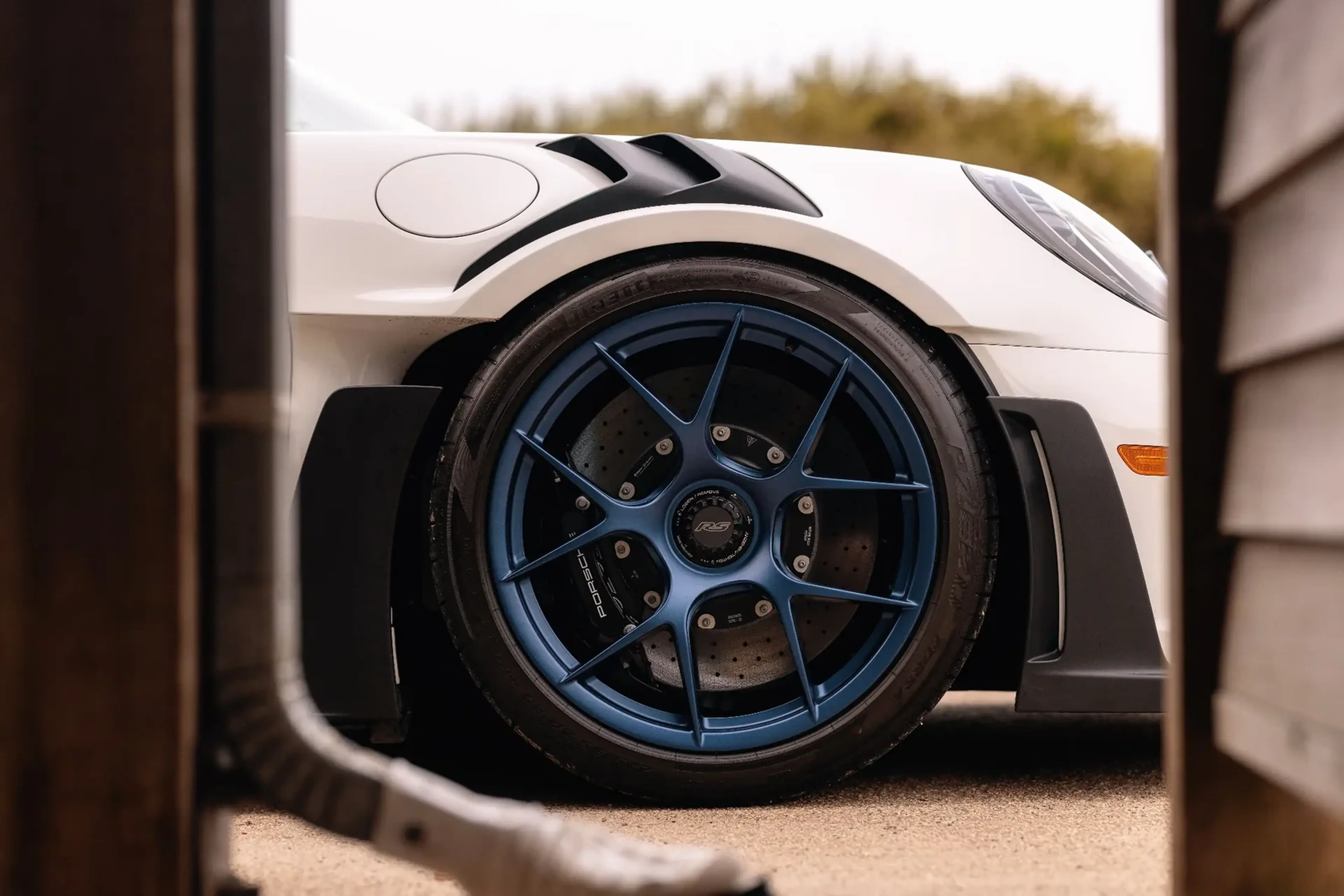 Close-up of a blue alloy wheel on a white sports car, viewed between wooden structures, with detailed brake system visible.