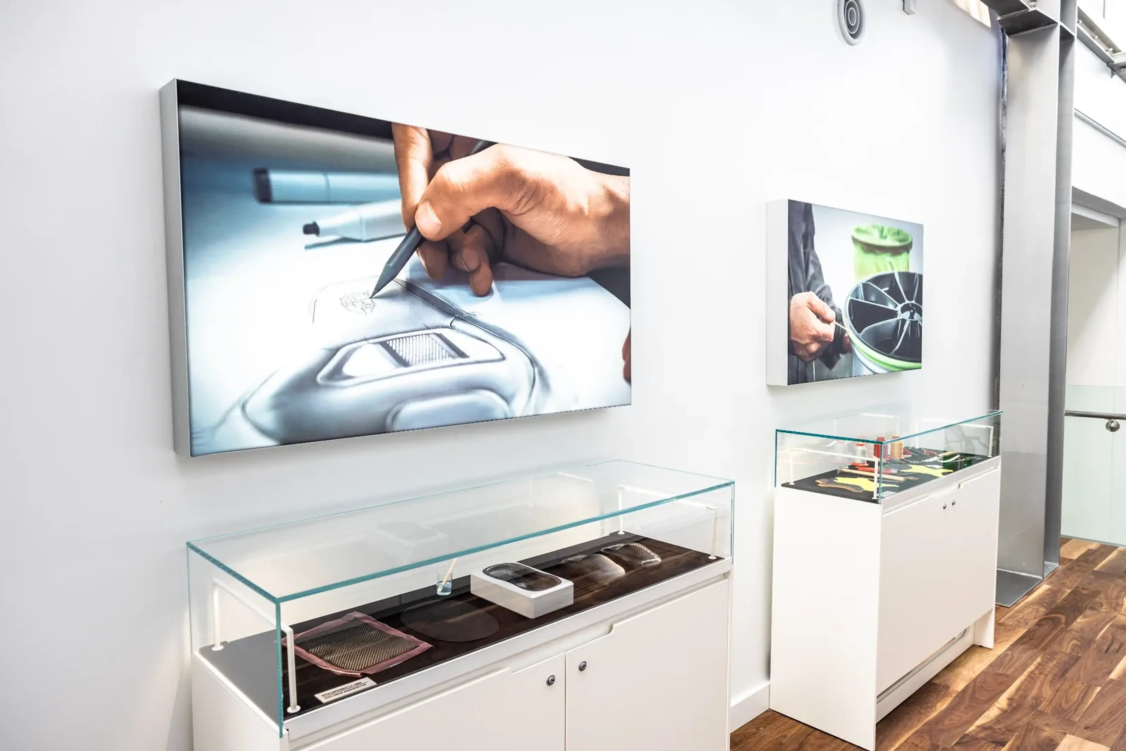 Two framed photos on a wall show detailed automotive design work, with hand sketches and parts, above display cases with various materials.