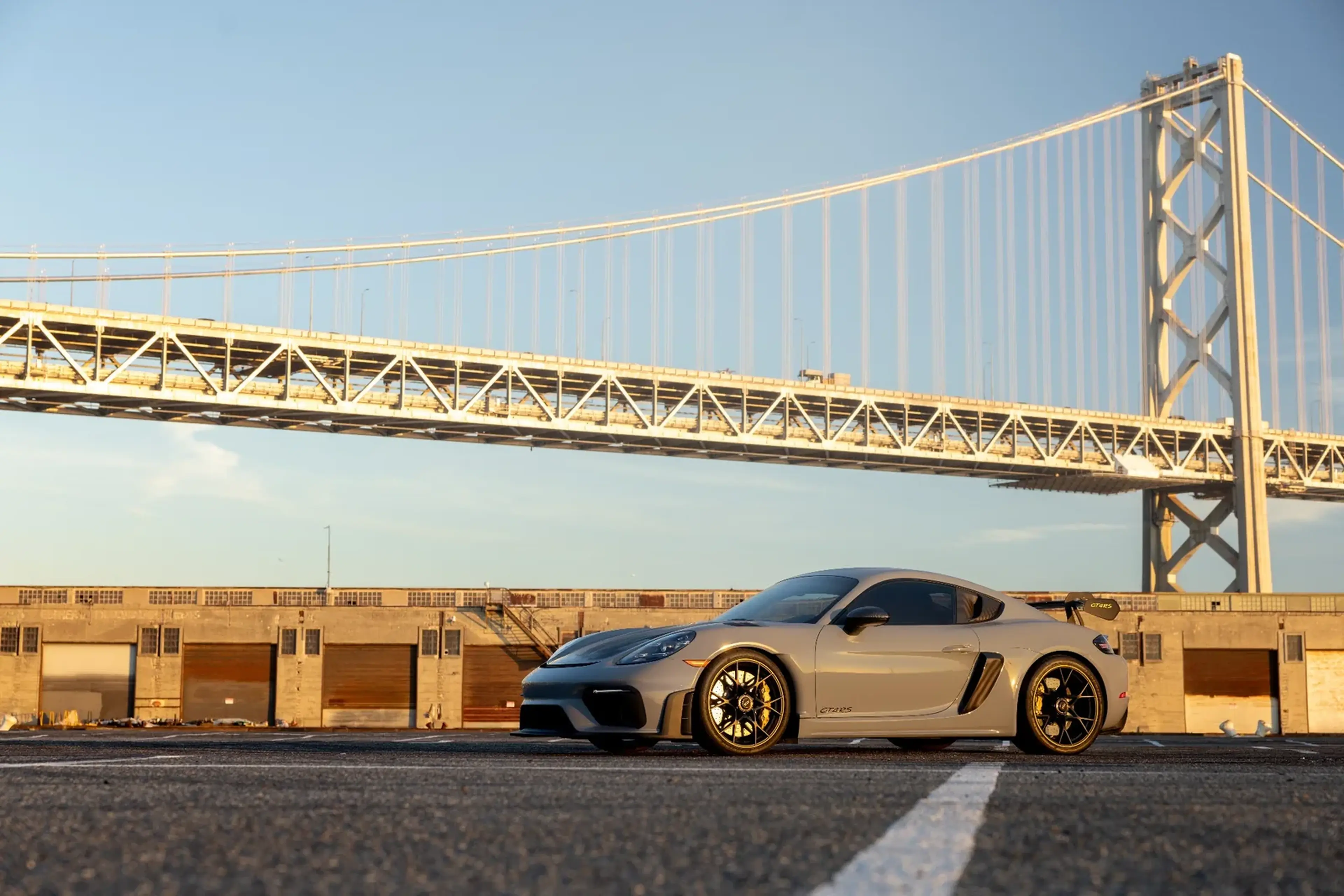Gray sports car parked in front of a large steel suspension bridge under a clear blue sky.