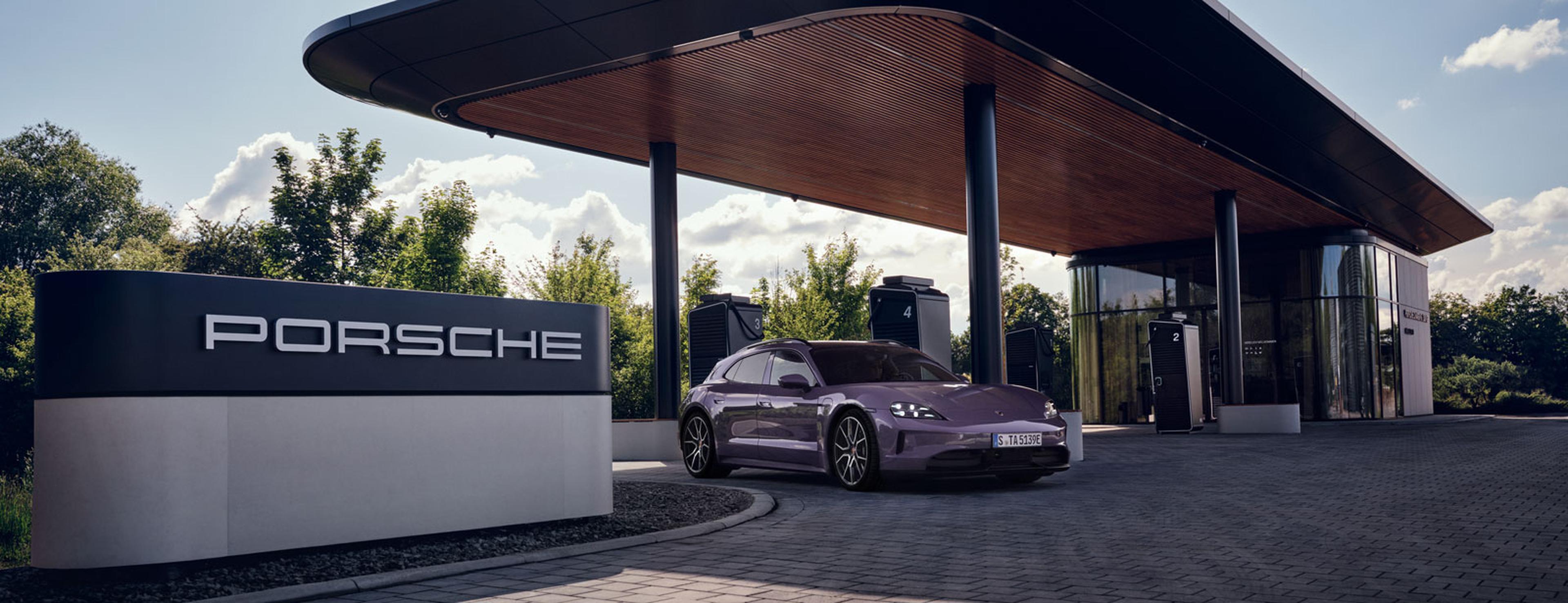 Ein violetter Sportwagen, geparkt an einer modernen Ladestation mit einem großen „Porsche“-Schriftzug, umgeben von Bäumen unter klarem Himmel.