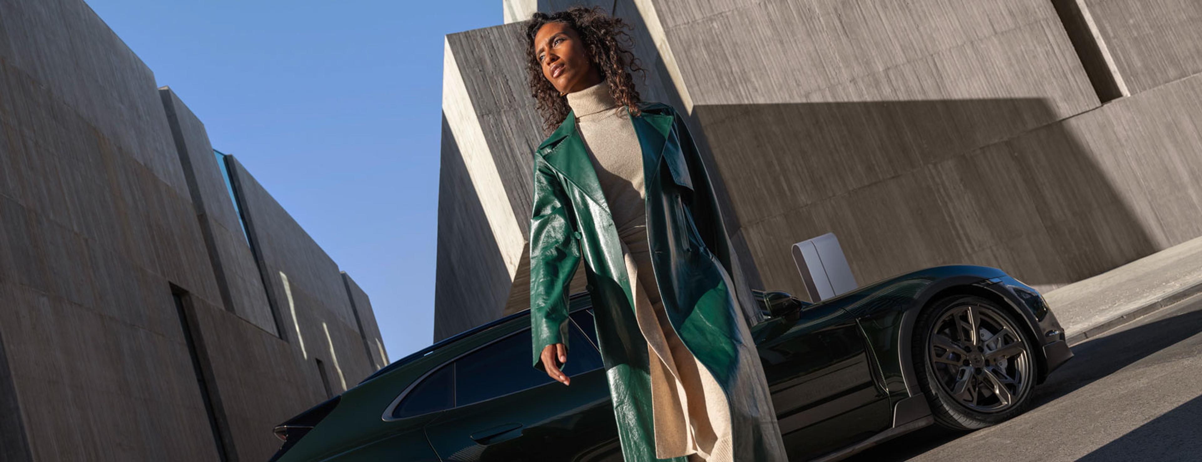 Eine Frau in einem grünen Ledermantel und einem beigen Kleid geht an einem dunklen Porsche vorbei; im Hintergrund sind moderne Betongebäude und ein klarer, blauer Himmel zu sehen.