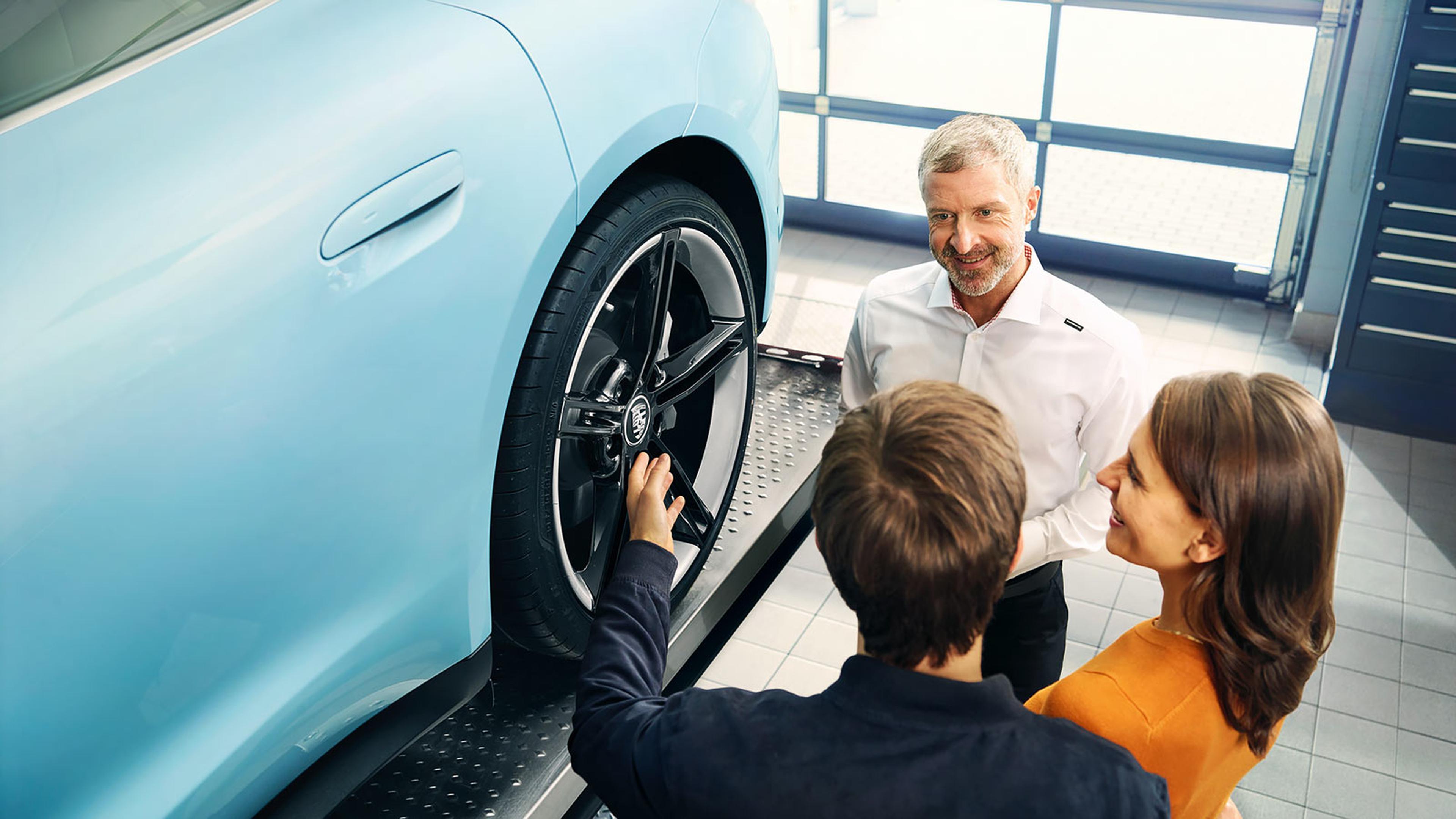 People discussing and inspecting a blue car's wheel on a lift in a garage setting.
