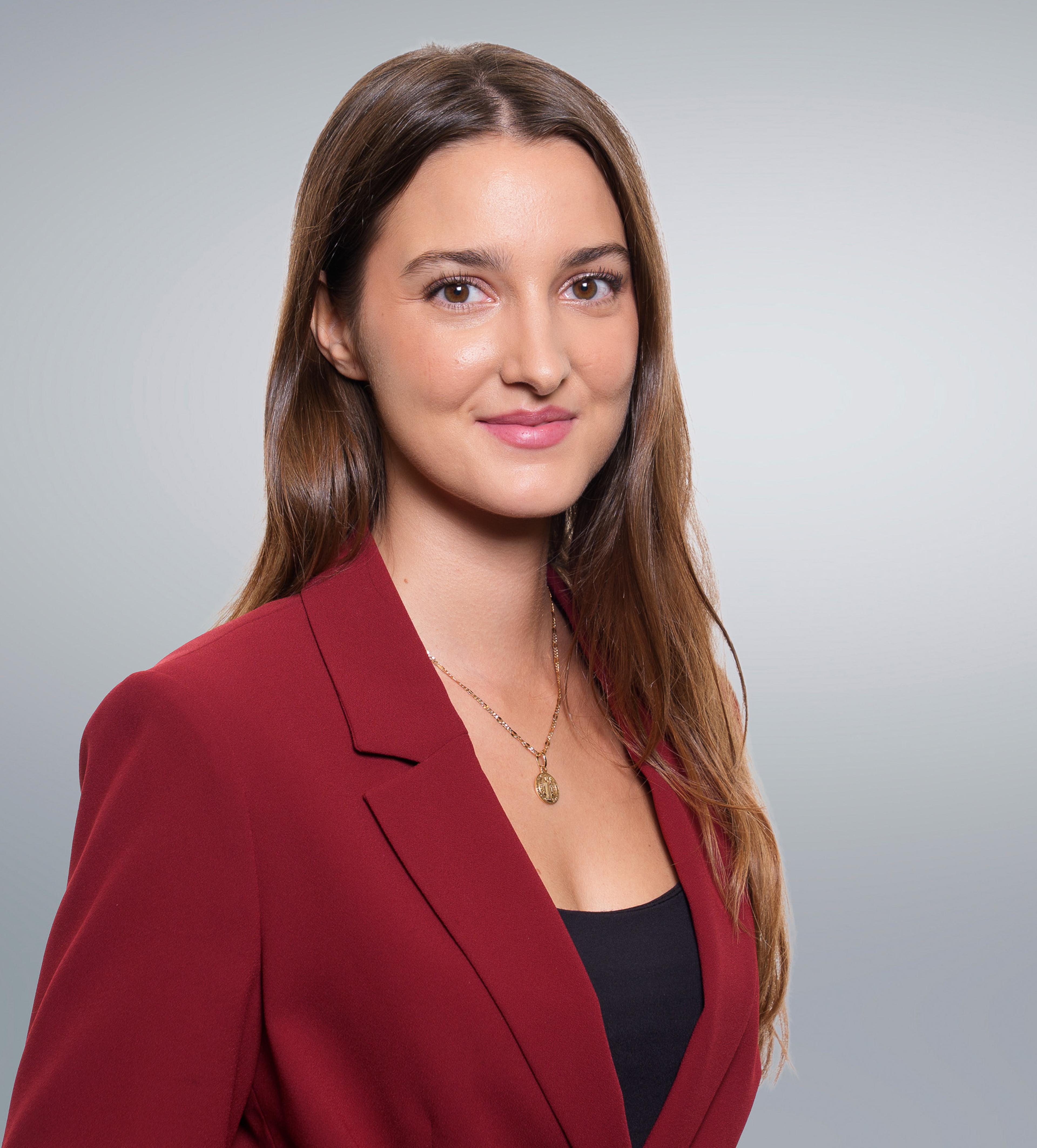 A woman with long brown hair in a red blazer and black top, smiling slightly against a plain, light gray background.