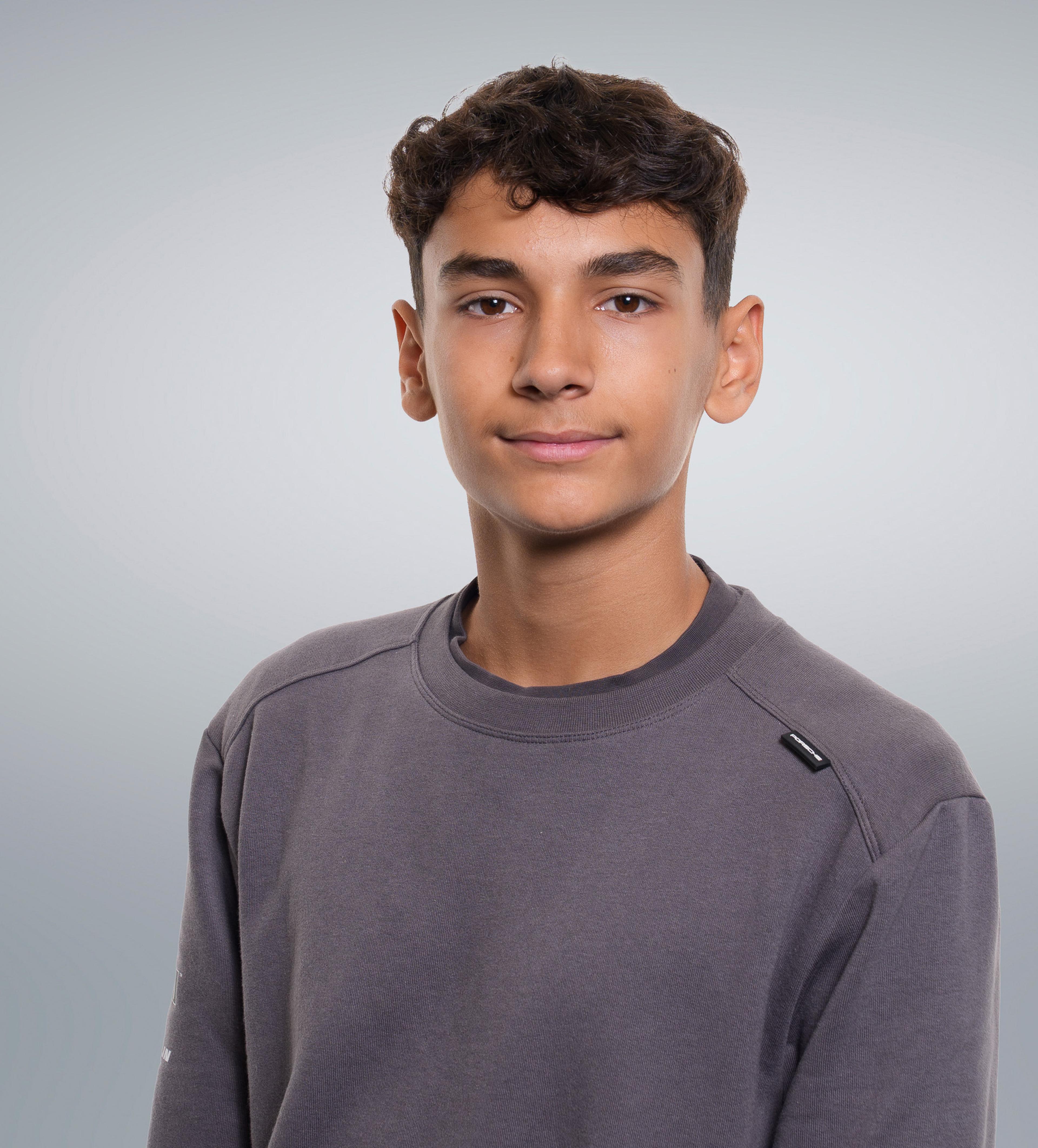 Teen boy with short, curly hair wearing a gray sweater, standing against a gray background.