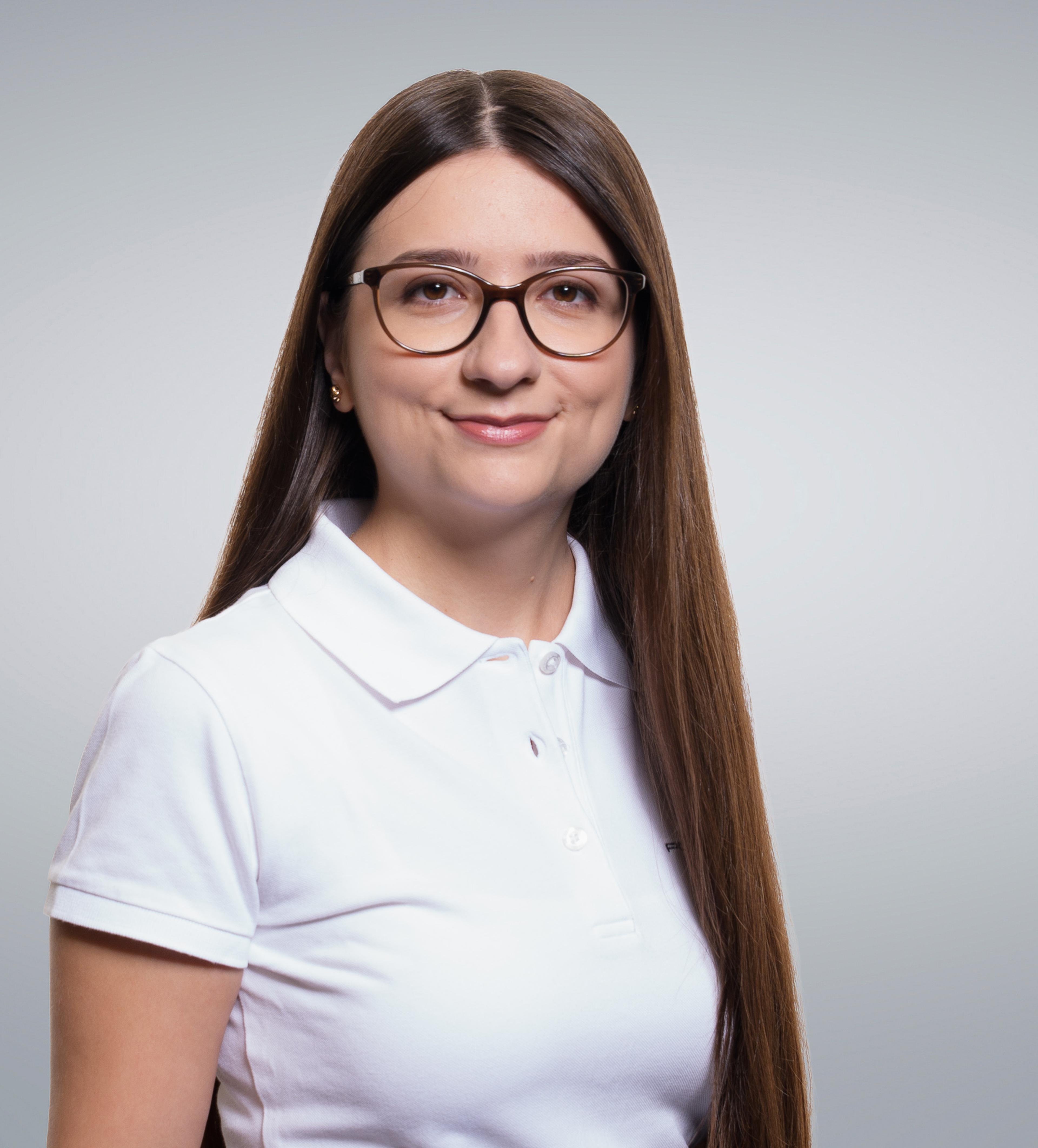 A woman with long brown hair and glasses smiles while wearing a white polo shirt, posed against a plain background.