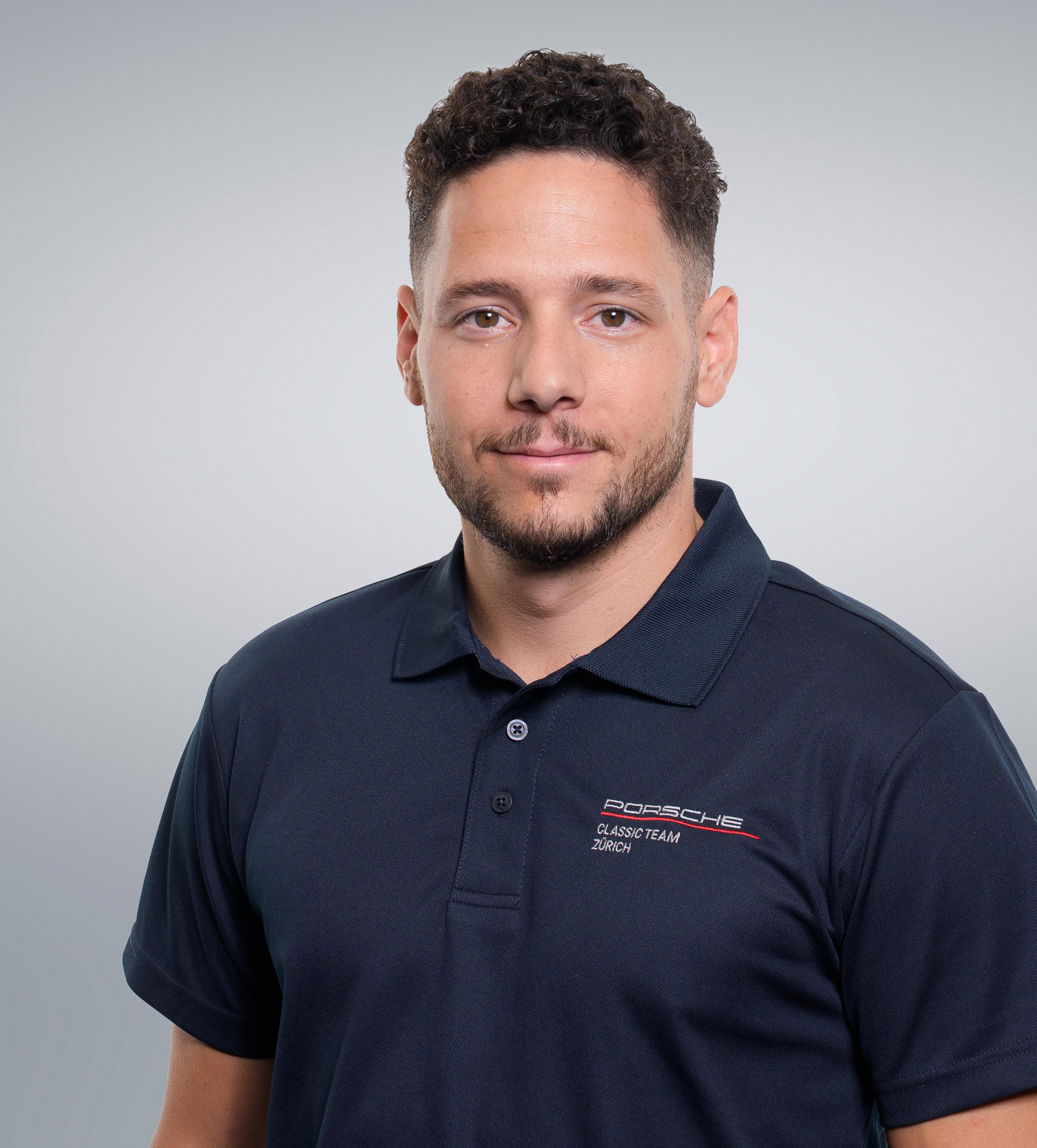 Man with curly hair in a navy blue polo shirt with "Porsche Classic Team Zürich" logo, standing against a plain background.