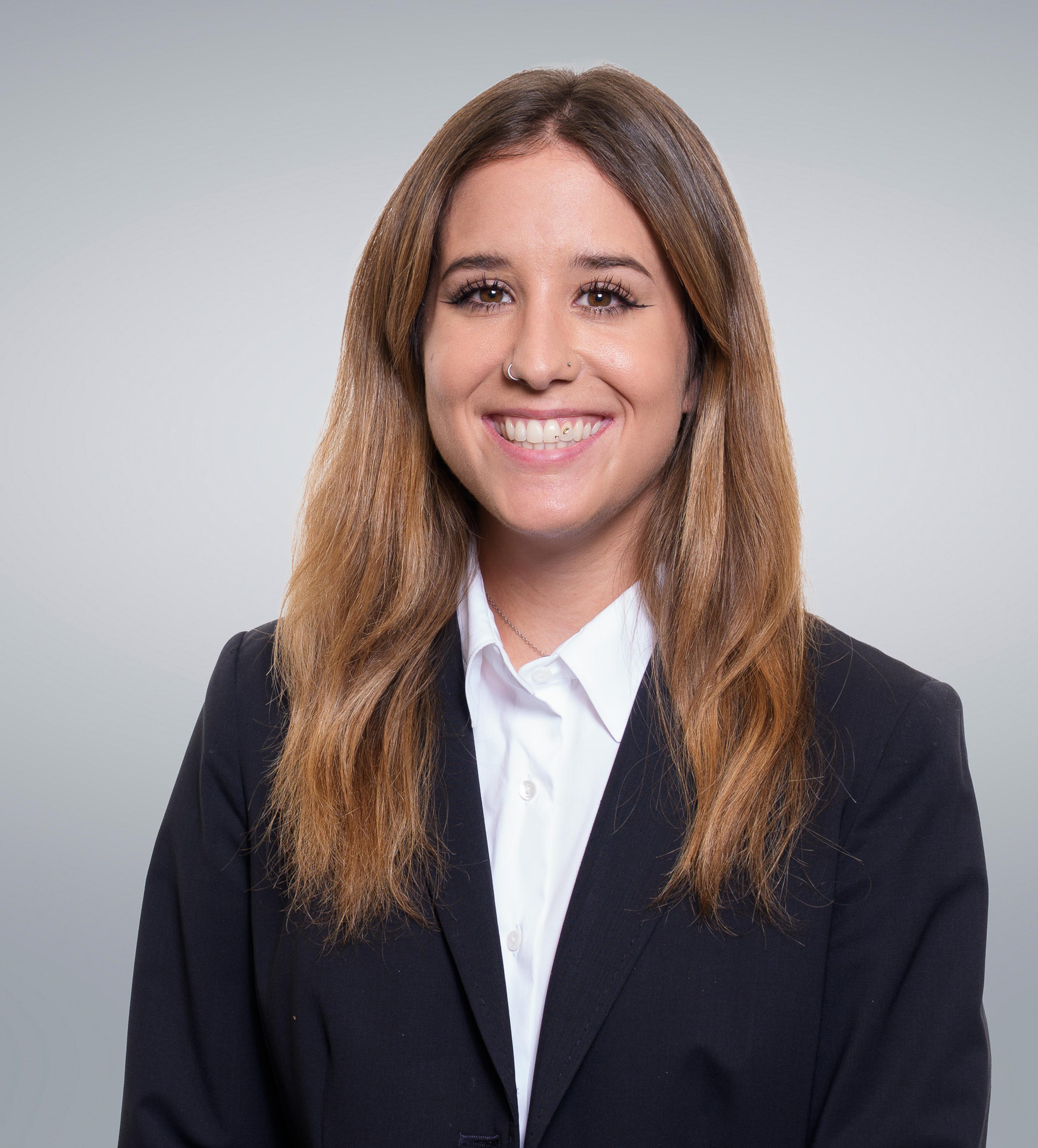 A smiling woman in a black blazer and white shirt, standing against a light gray background.