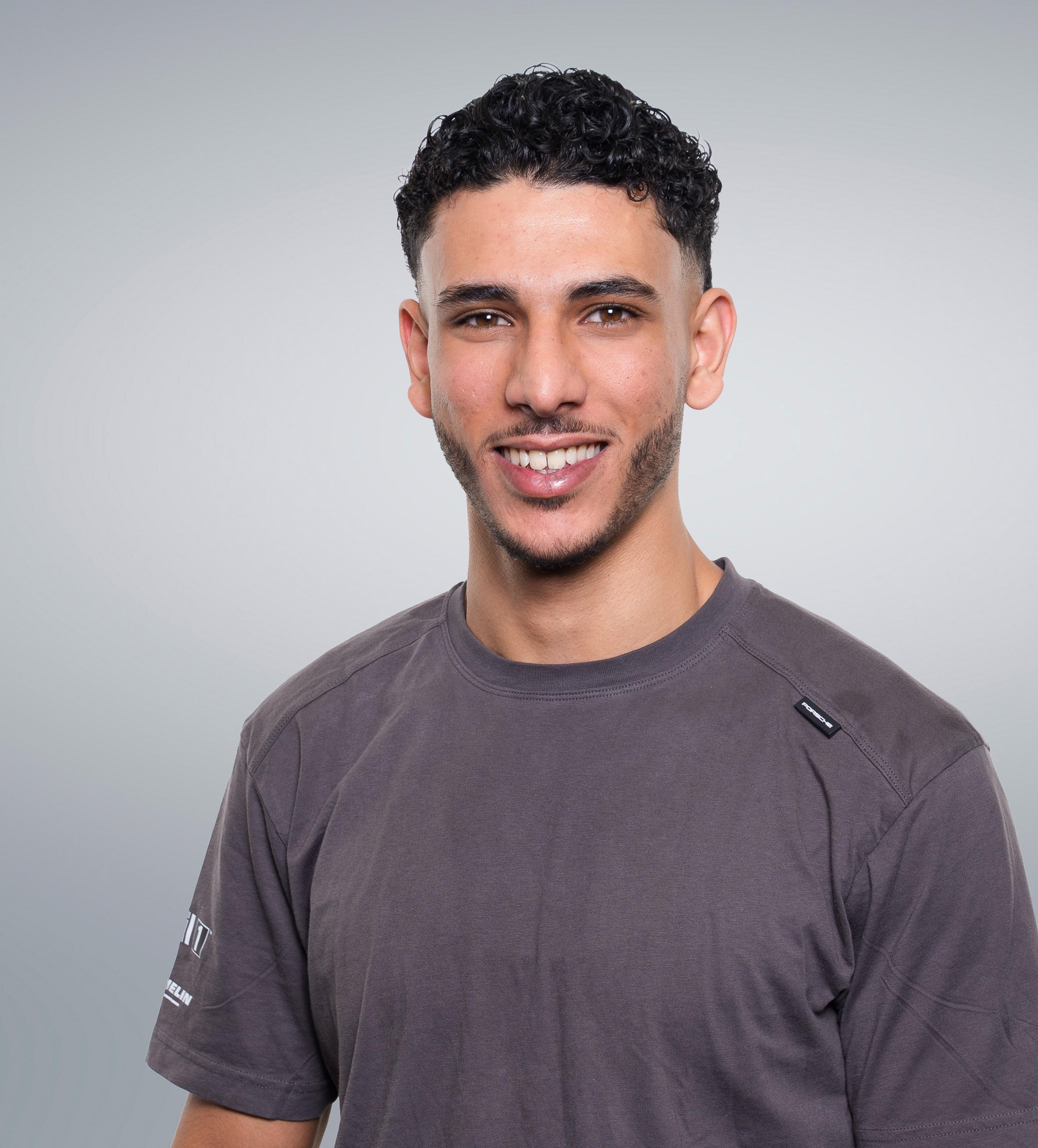 Smiling man with short curly hair and a beard, wearing a gray t-shirt, against a plain gray background.