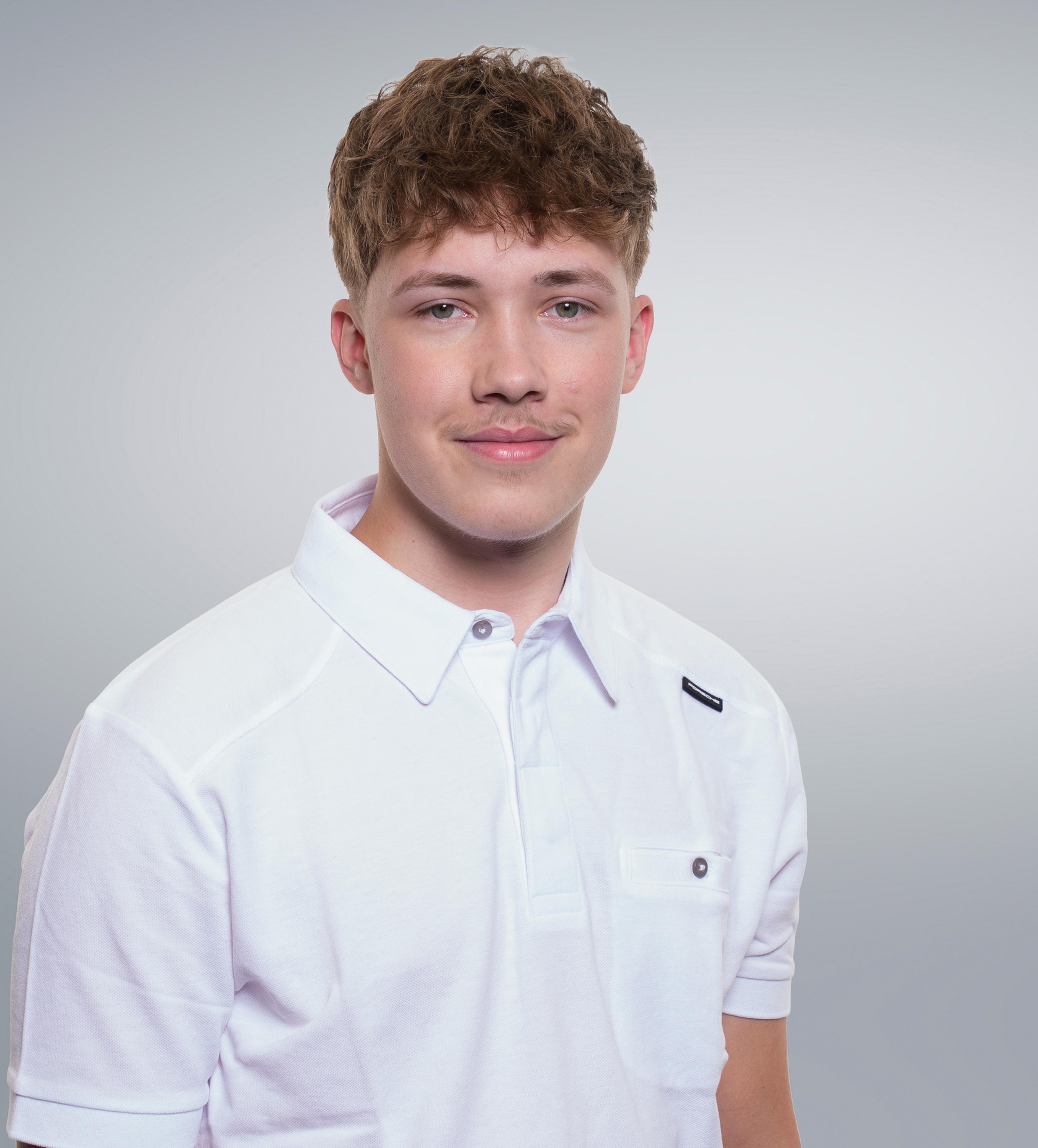 Young man with short curly hair and a subtle mustache wearing a white polo shirt, smiling gently against a neutral background.
