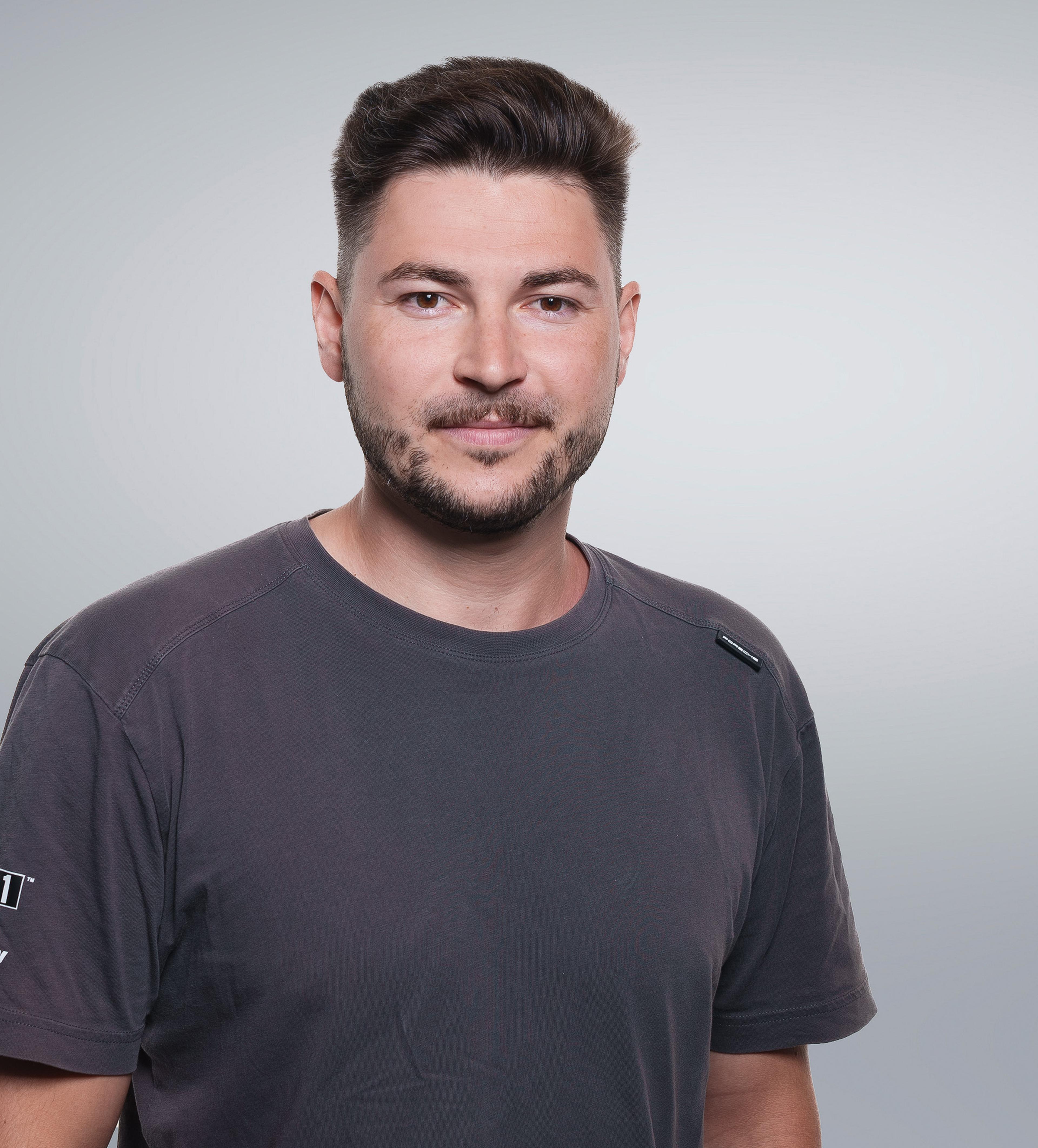 A man with short dark hair and a beard smiles slightly while wearing a gray T-shirt against a plain white background.
