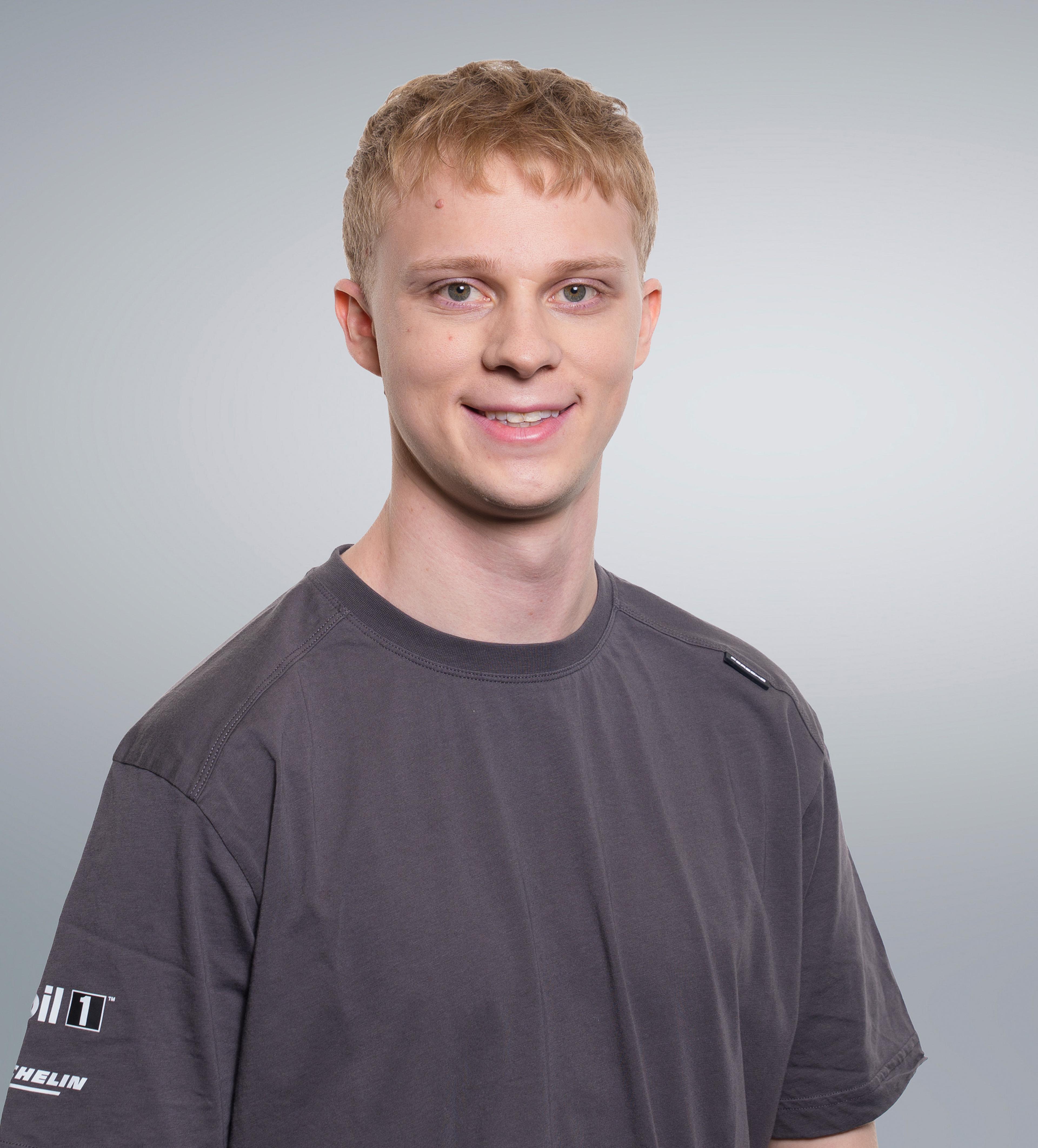 Young man with short blond hair wearing a dark gray T-shirt, smiling in front of a plain gray background.