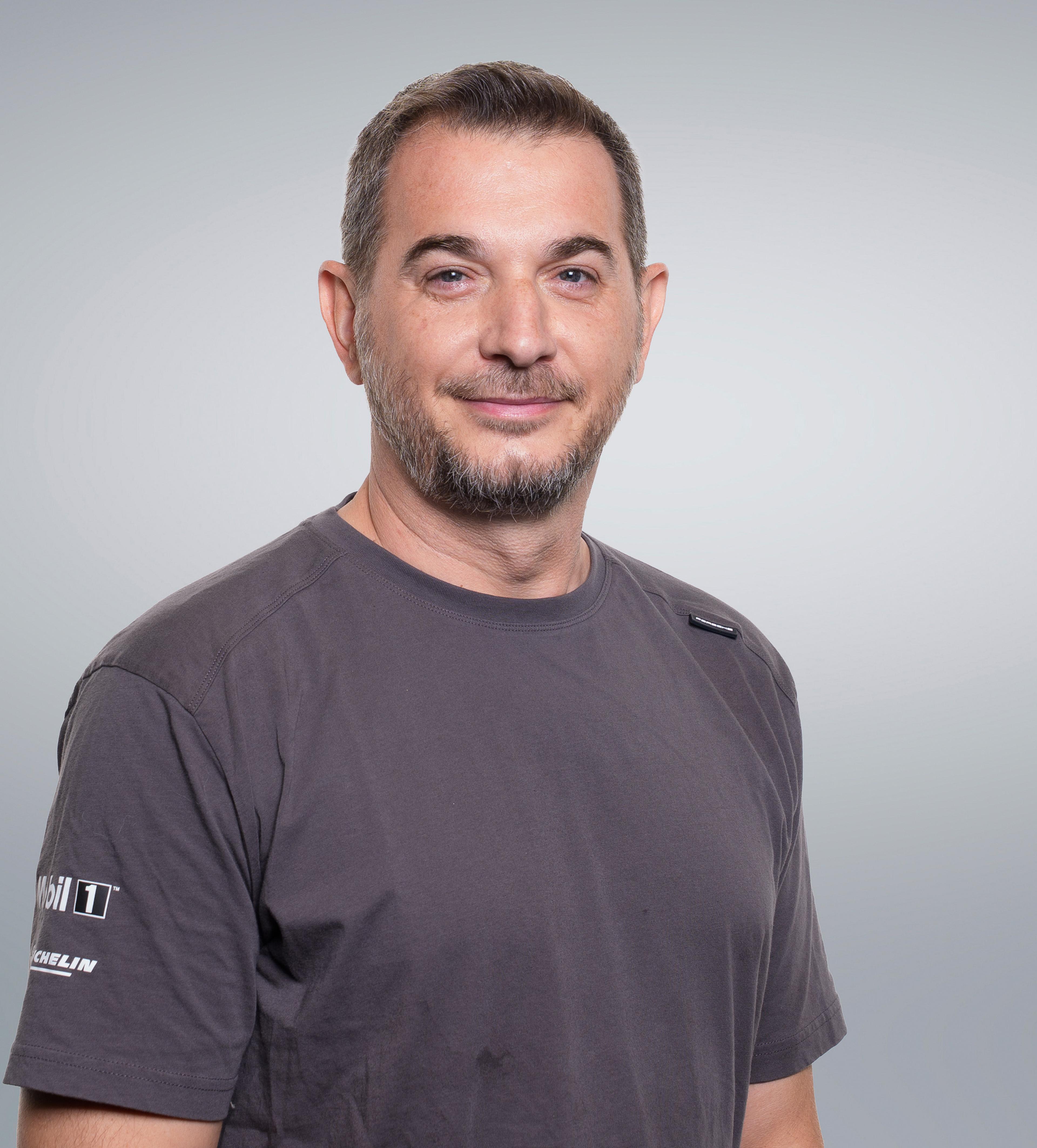 A man with short hair and a beard smiling, wearing a gray T-shirt with logos on the sleeves, standing against a light gray background.