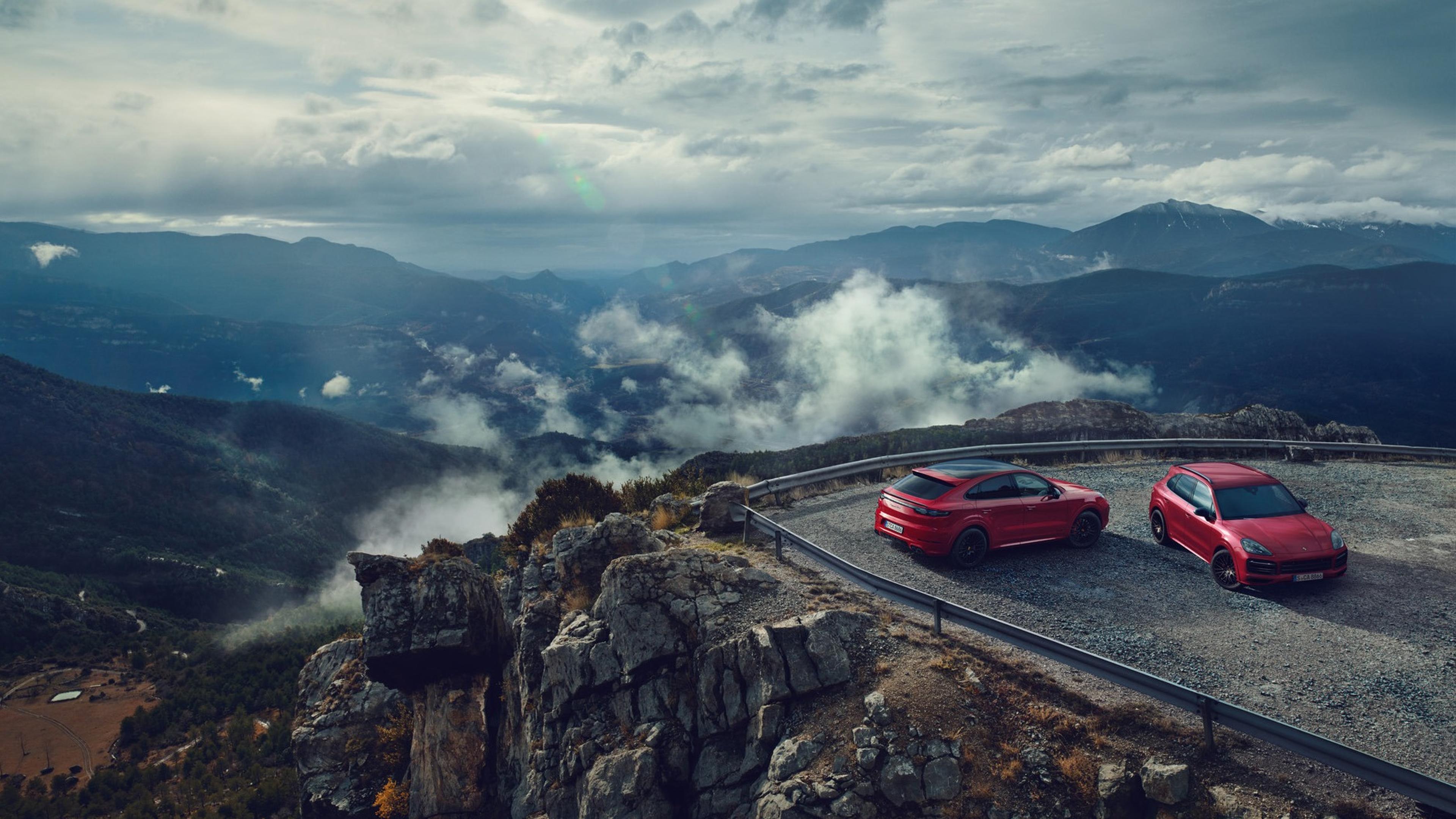 Two Cayenne coupe GTS 's parked at a mountain viewpoint overlooking cloudy, scenic valleys under a dramatic sky.