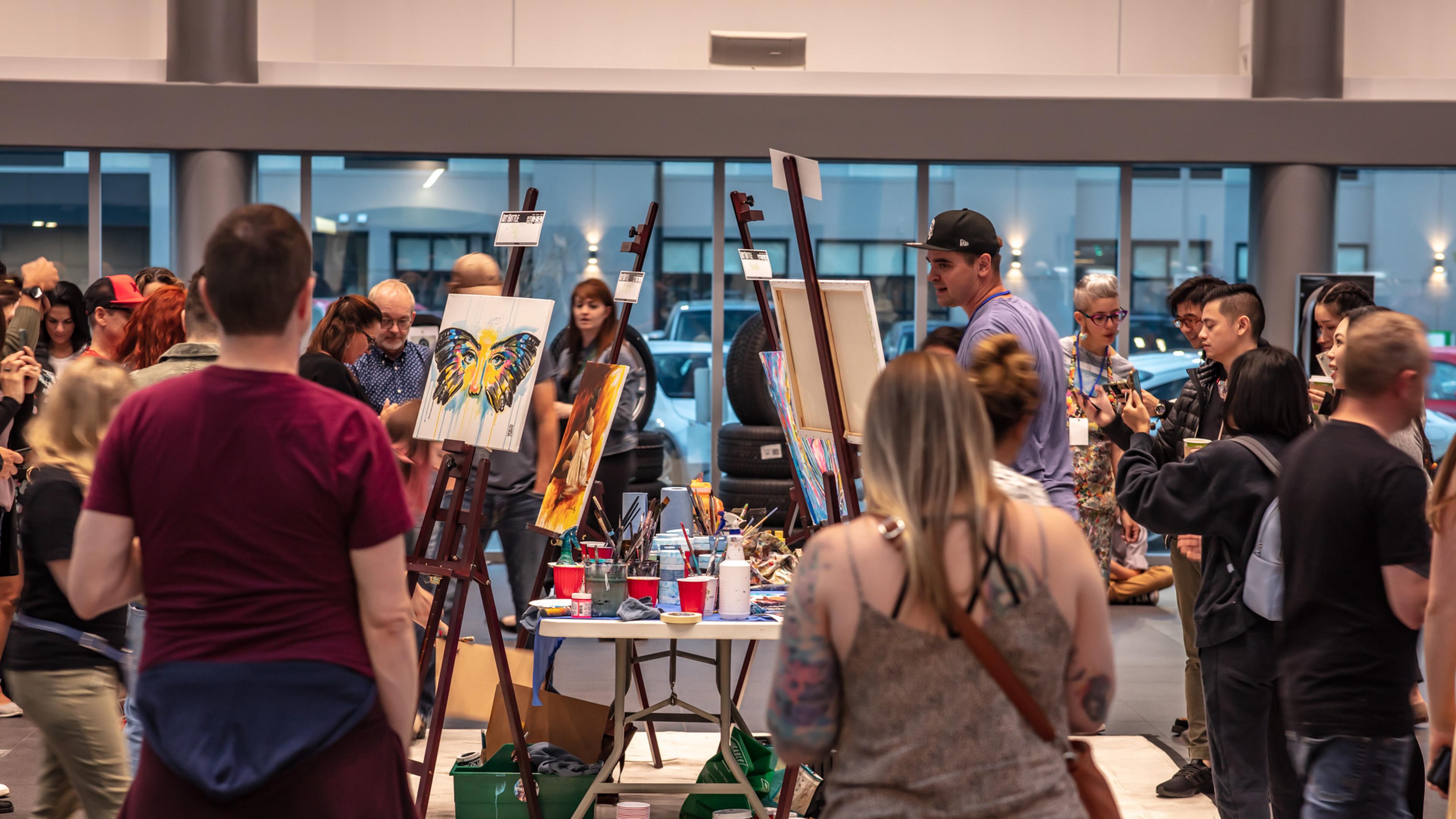A crowd gathers at Porsche Centre Calgary around easels displaying vibrant artwork in a well-lit, modern gallery space, engaging with the art and each other.