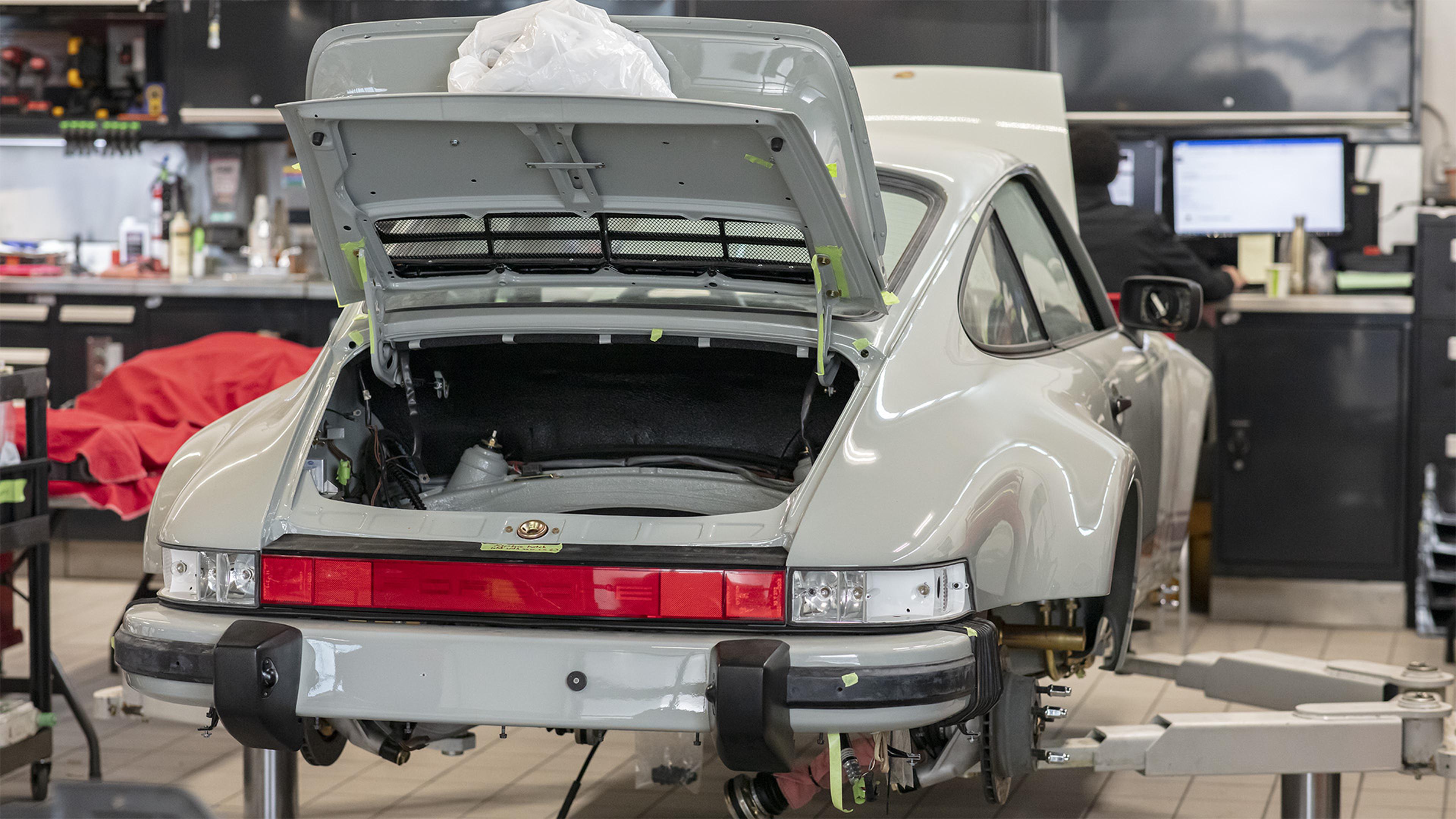 Wide body 964 in chalk from cosmos collisions, rear engine compartment open, undergoing maintenance in a garage. Tools and equipment are visible in the background.