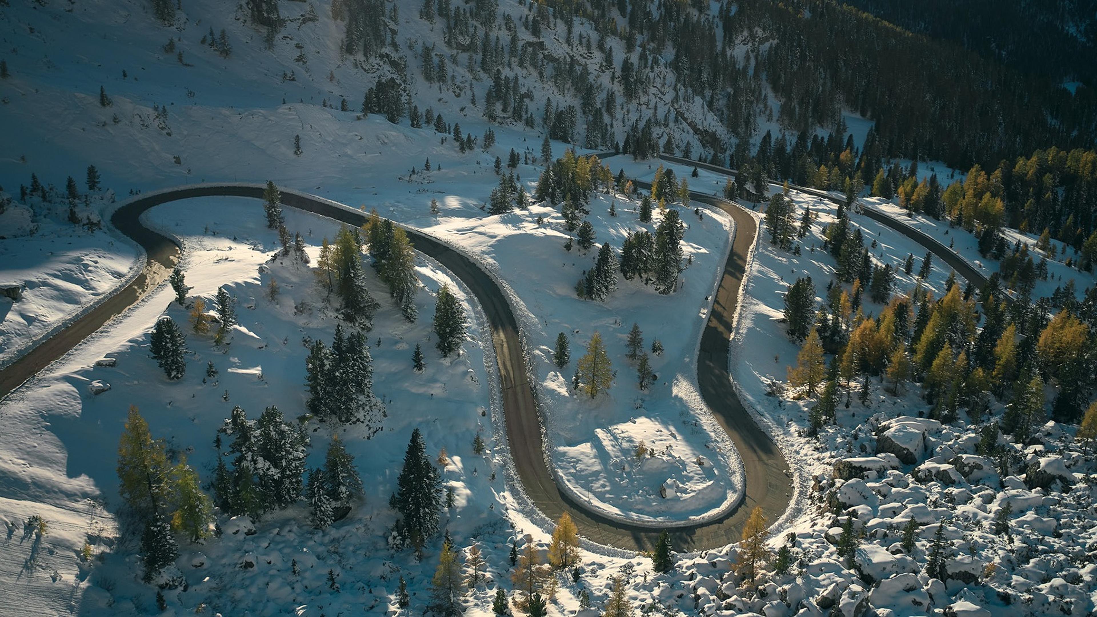 A winding road curves through a snowy, mountainous landscape dotted with evergreen trees under a clear sky.