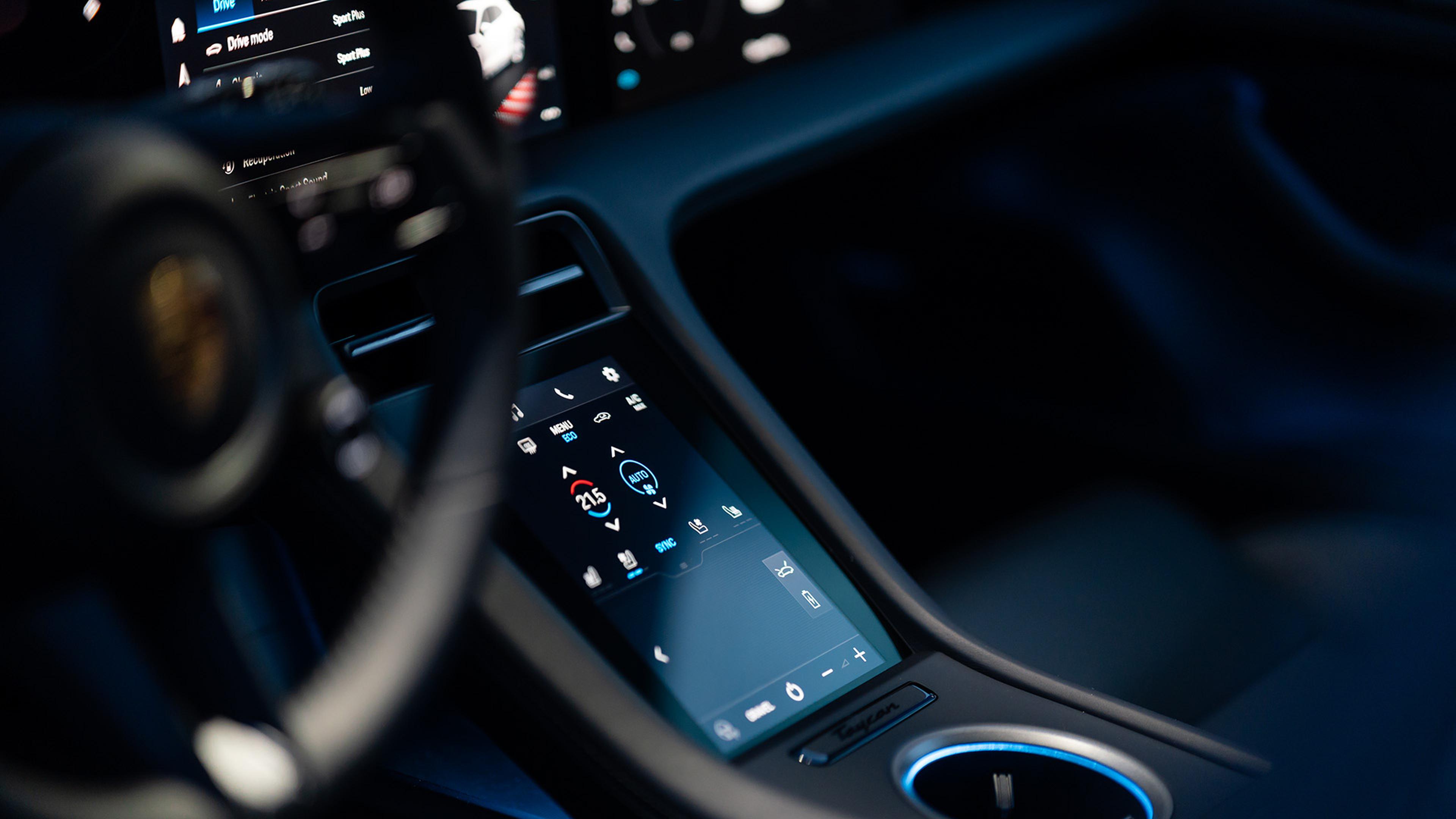 Taycan Close-up of a car's digital touchscreen dashboard, displaying climate control settings and driving modes, with part of the steering wheel visible.
