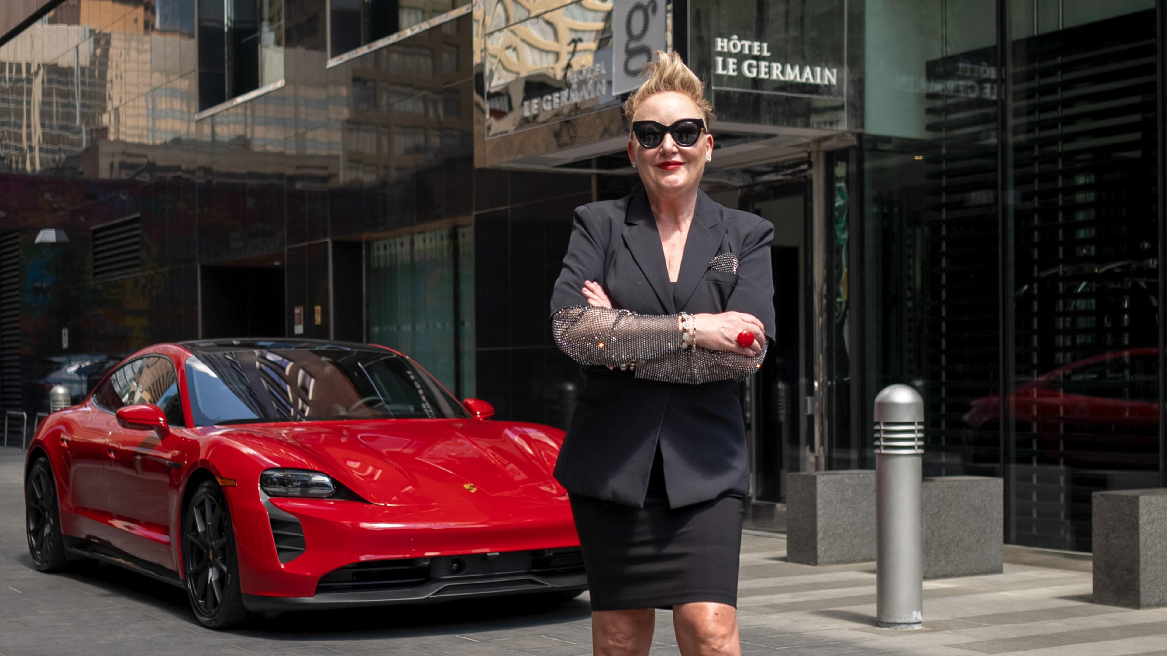 A confident person in a stylish black outfit stands in front of a sleek Porsche Taycan GTS in Carmine Red outside Hôtel Le Germain.