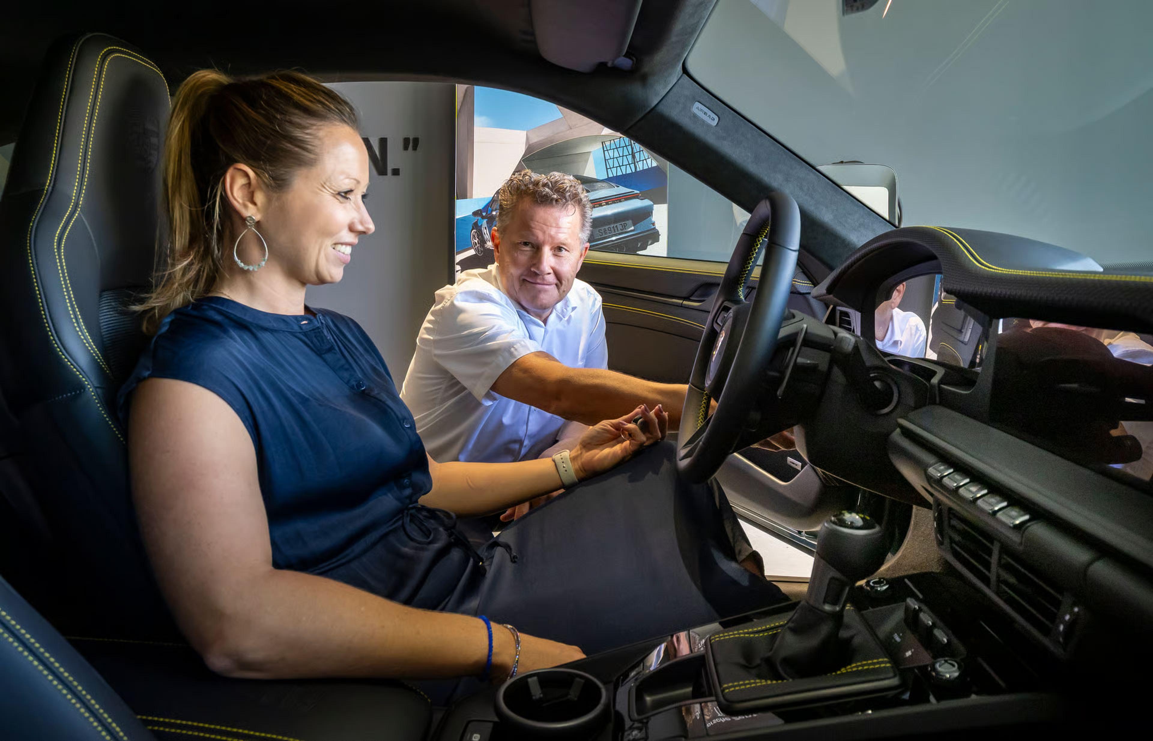 Eine Frau sitzt lächelnd auf dem Fahrersitz eines Autos, während ein Mann die Funktionen im Fahrzeuginneren erklärt.