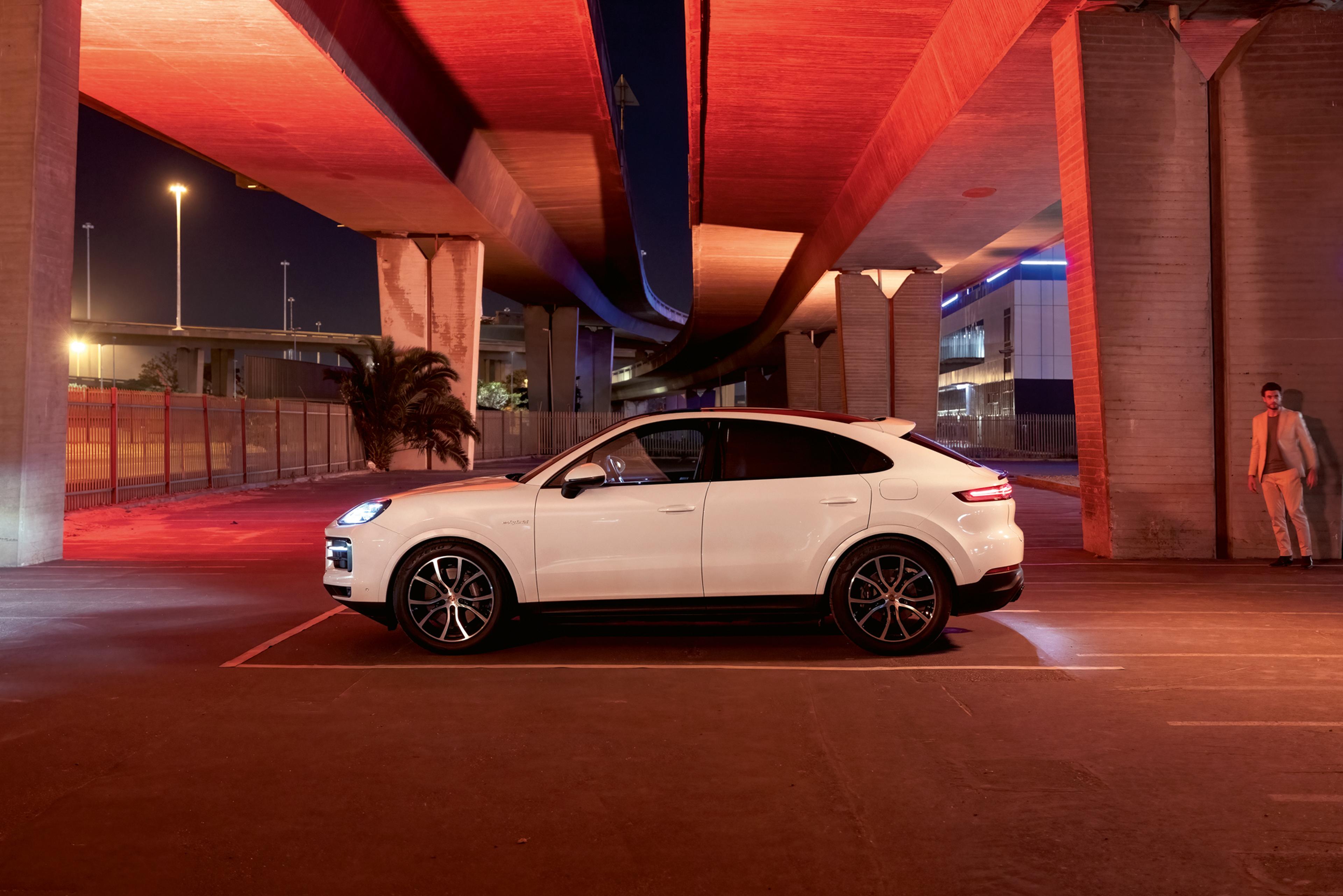 Ein weißer Porsche Macan, der nachts unter einer Autobahnbrücke geparkt ist, wobei rotes Licht die Szene beleuchtet. Im Hintergrund steht eine Person.