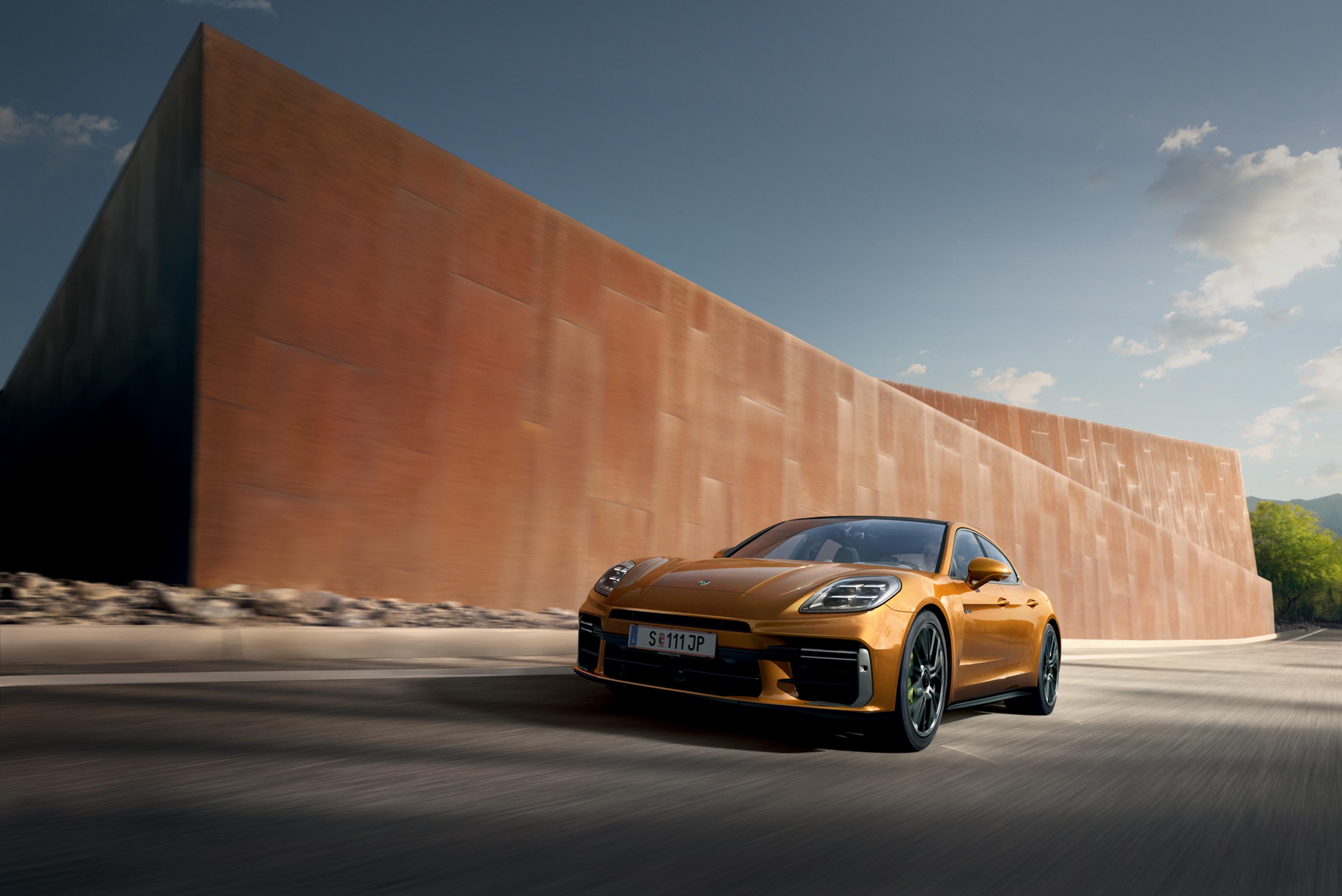 Ein schnittiger, orangefarbener Porsche Panamera fährt auf einer kurvigen Straße neben einer großen, rostfarbenen Metallkonstruktion unter einem klaren blauen Himmel.