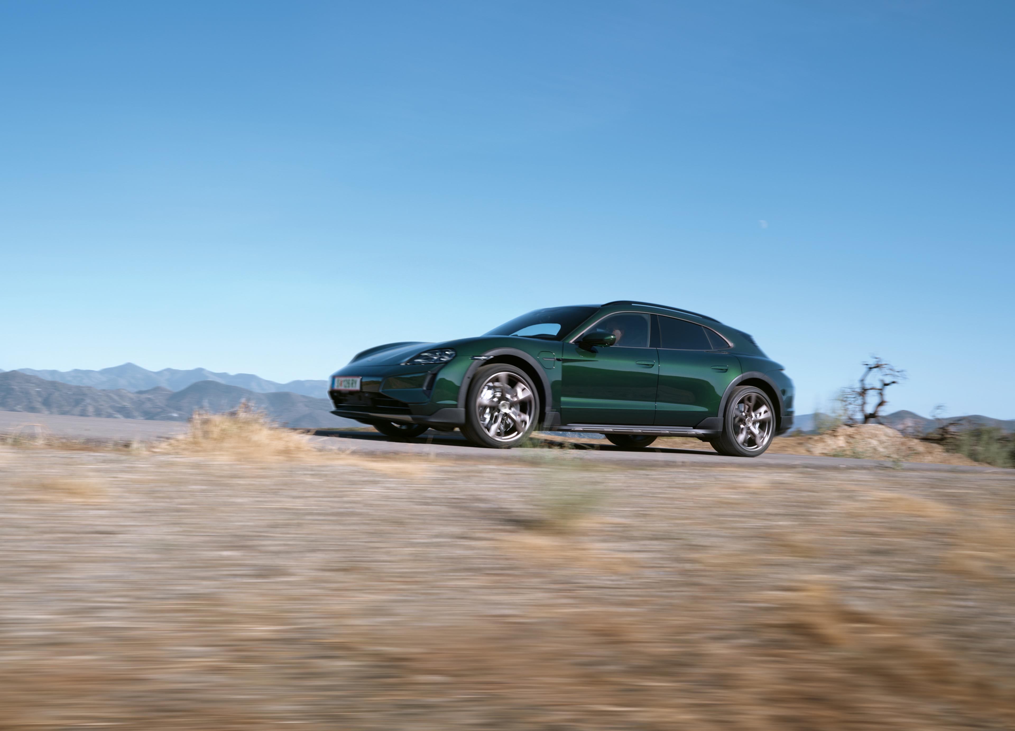 Ein grüner Porsche Taycan fährt auf einer Landstraße; im Hintergrund sind Berge und ein klarer, blauer Himmel zu sehen.
