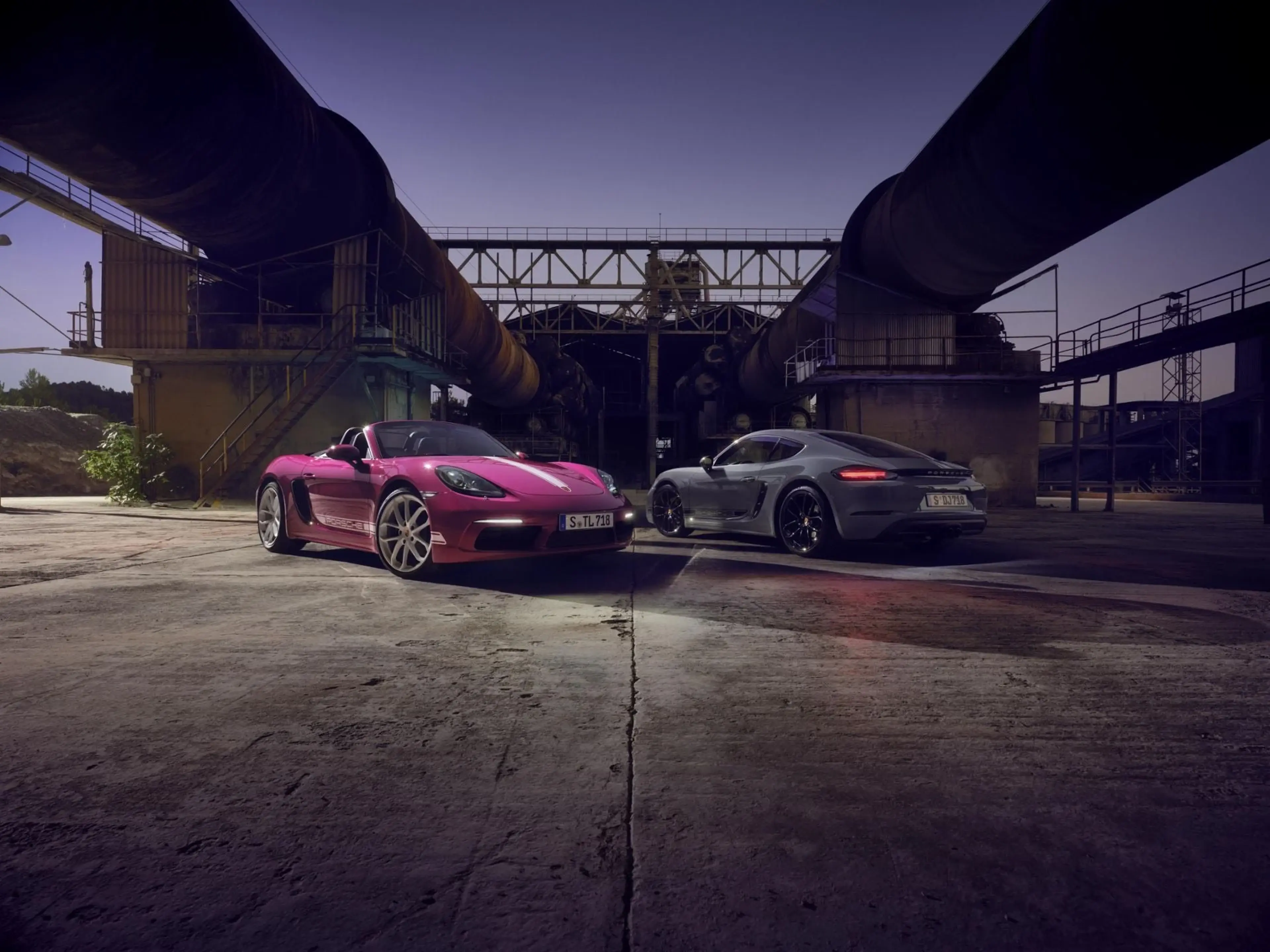 Porsche 718 Cayman models parked at dusk near an industrial bridge