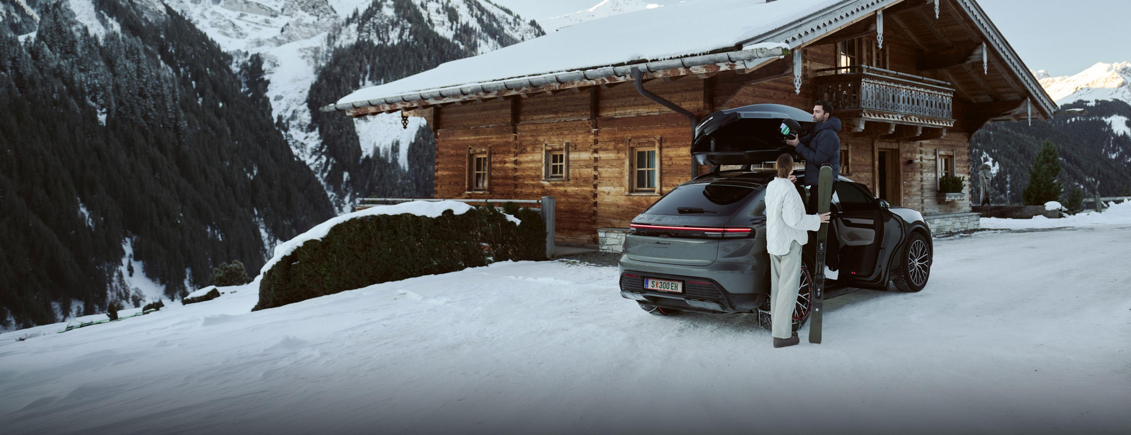 Ein Paar lädt neben einer Holzhütte in einer verschneiten Berglandschaft Skiausrüstung in ein Auto.
