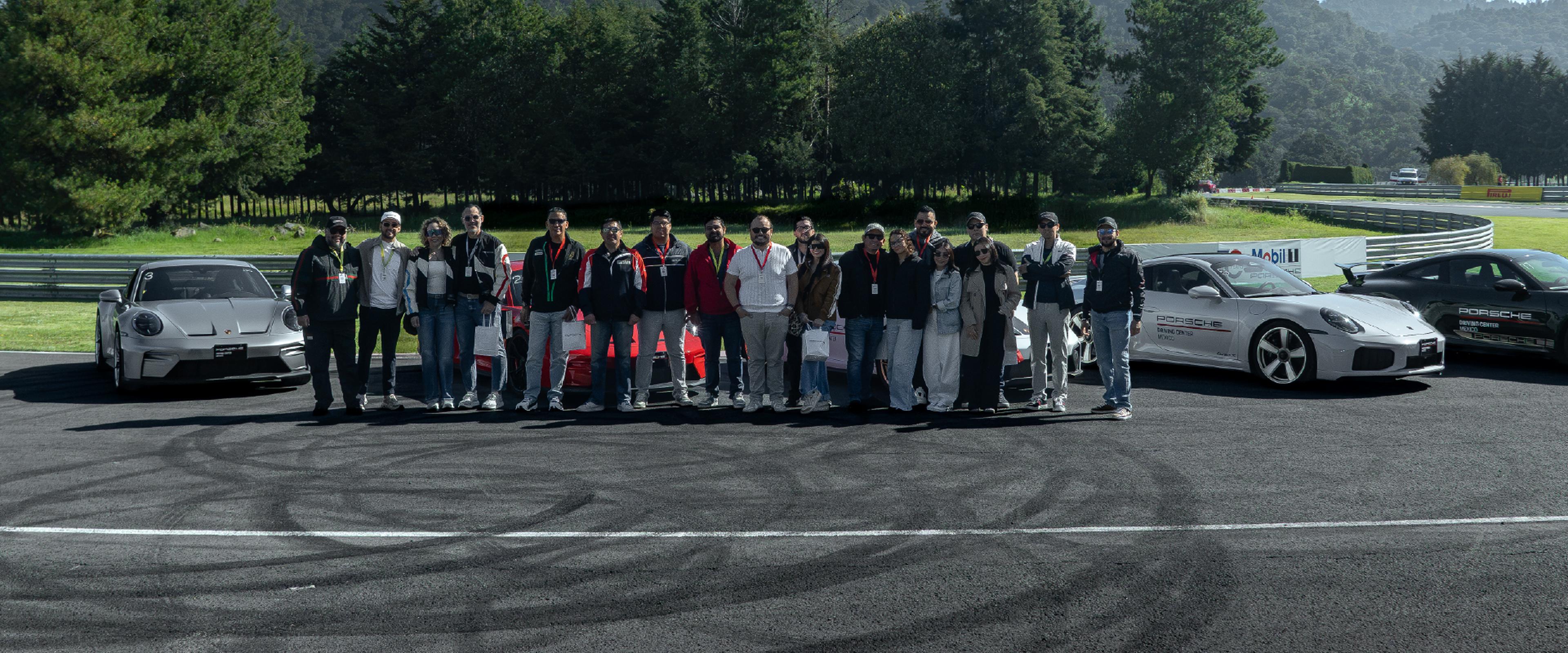 Un grupo de personas de pie en una pista de carreras, flanqueadas por varios coches deportivos Porsche, con árboles y una barrera al fondo.