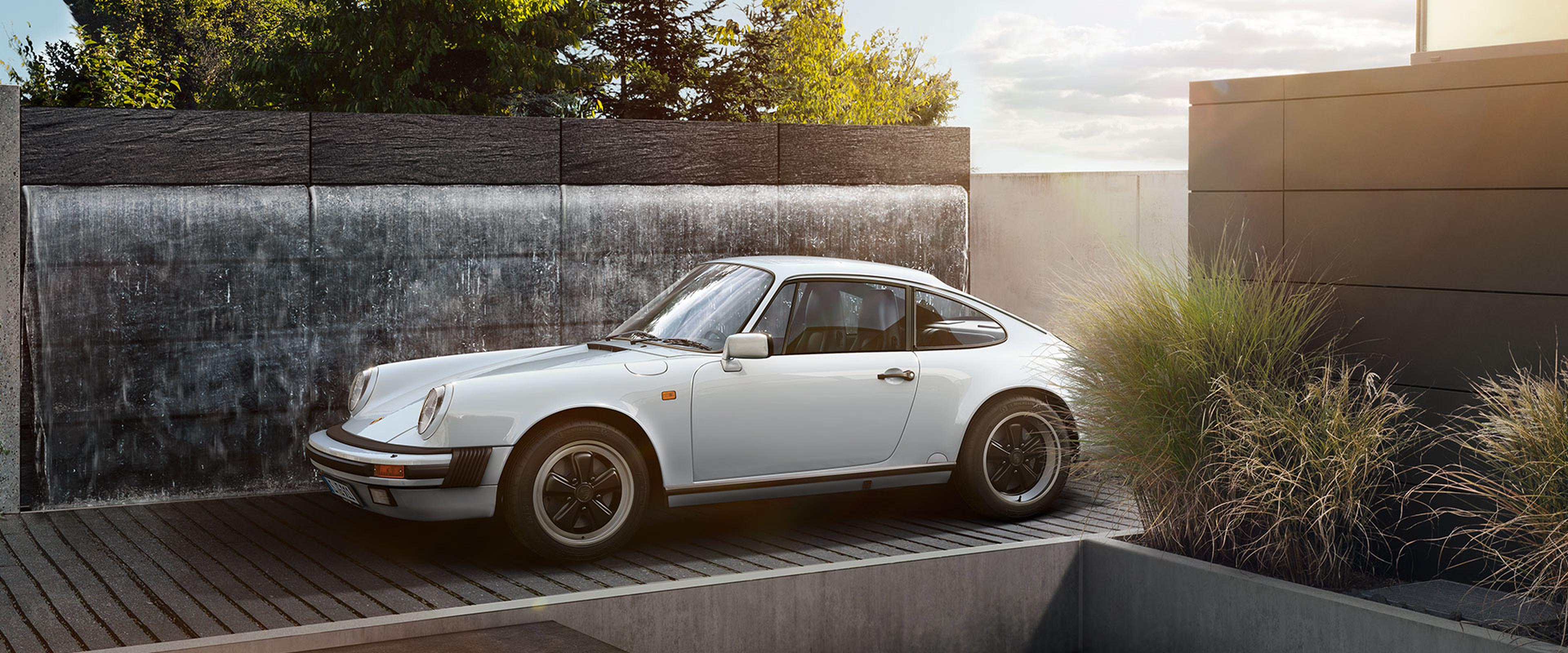 A classic white sports coupe parked on a modern wooden driveway, surrounded by landscaped greenery and sleek concrete walls under a bright sky.