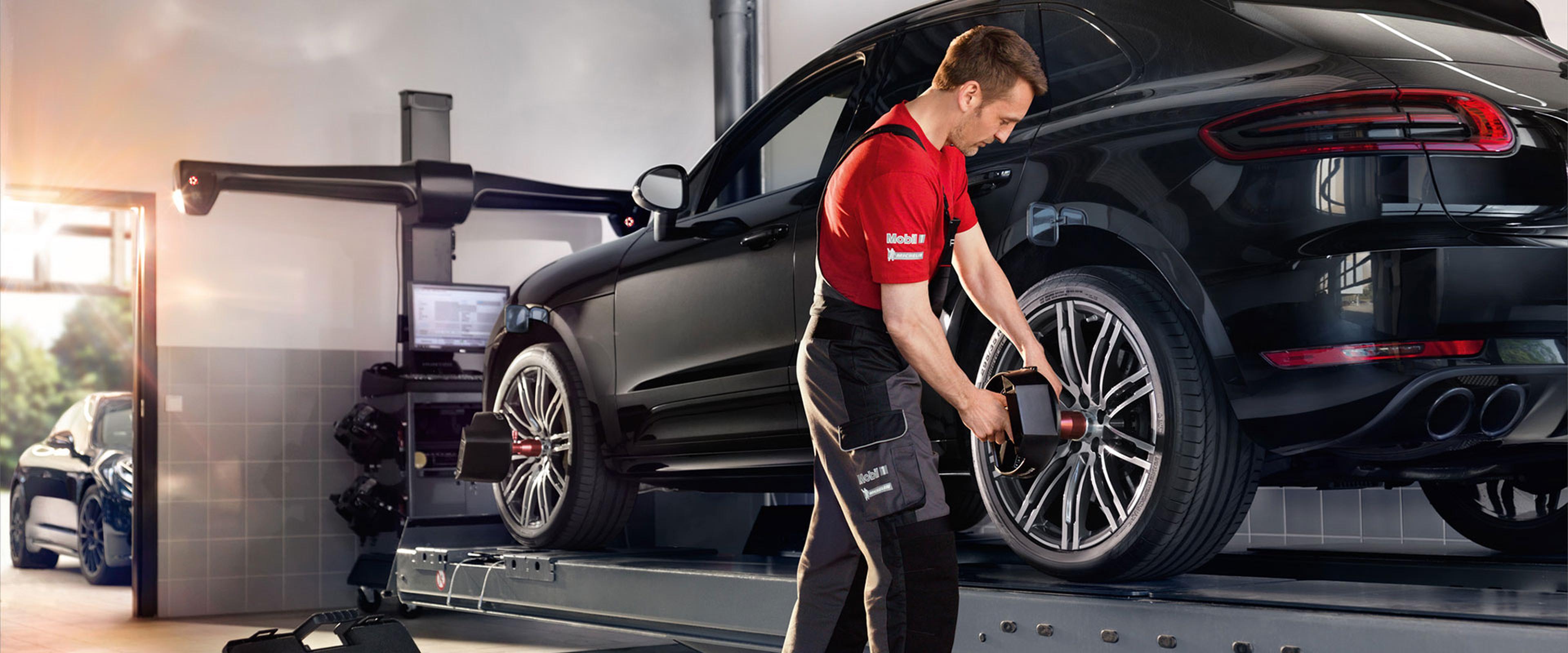 Mechanic in a red shirt and overalls working on a black car's tire in a garage with equipment and another car in the background.