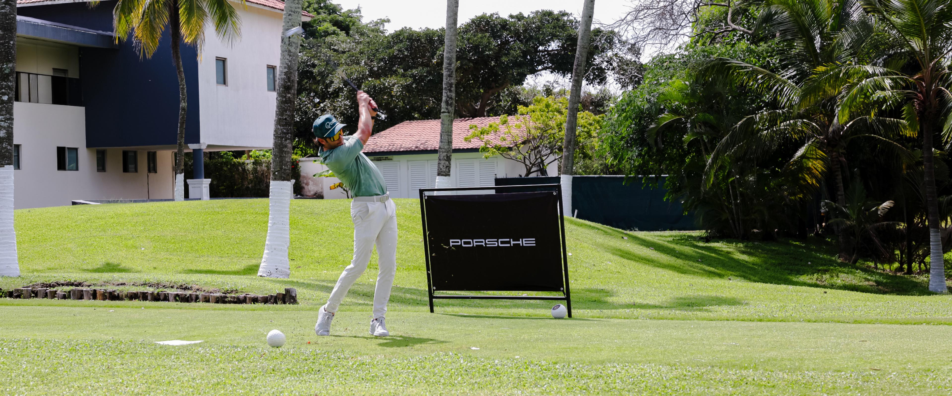 Un golfista a mitad de su swing en un campo soleado, rodeado de palmeras, con un cartel de Porsche al fondo.