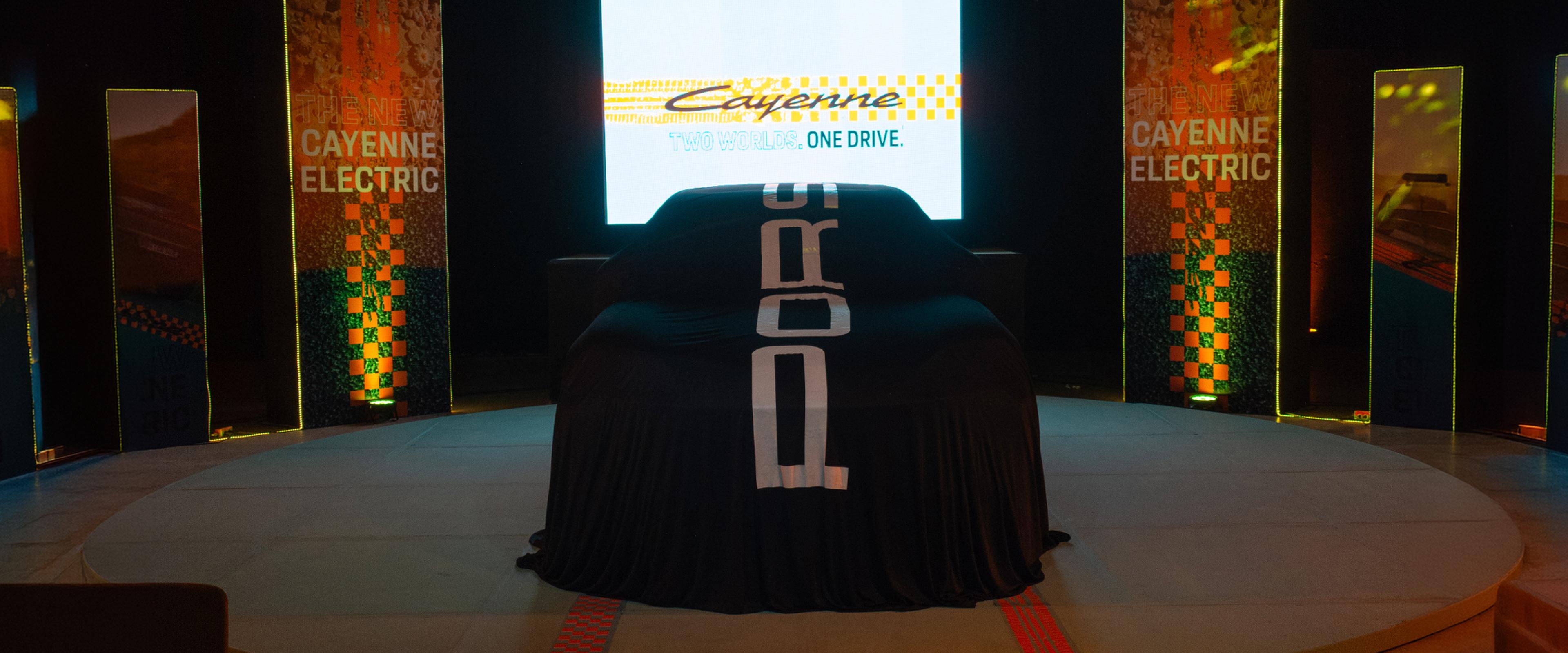 Automóvil cubierto sobre un escenario con un telón de fondo que dice «Cayenne Electric»; un evento de presentación de vehículo.