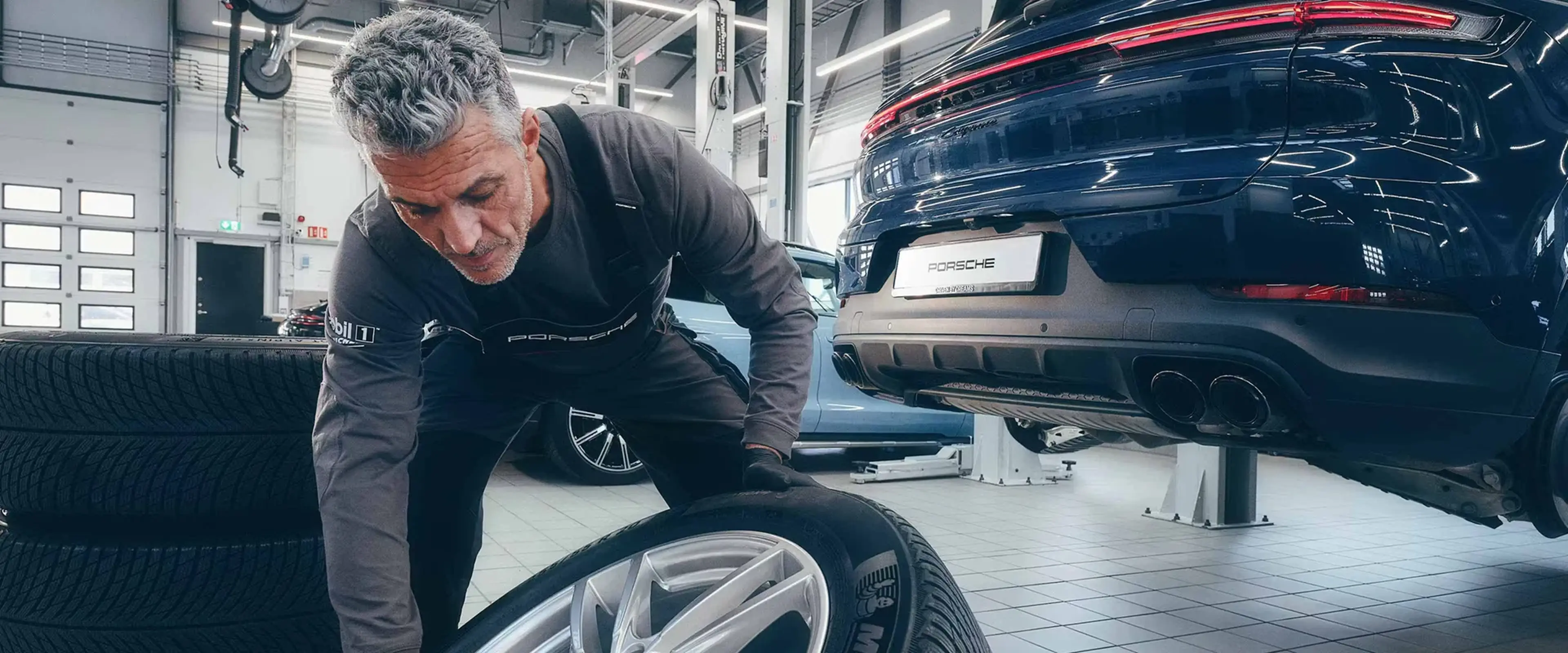 Man with grey hair and beard in a garage, changing a car tire. A blue car is raised on a hydraulic lift in the background.