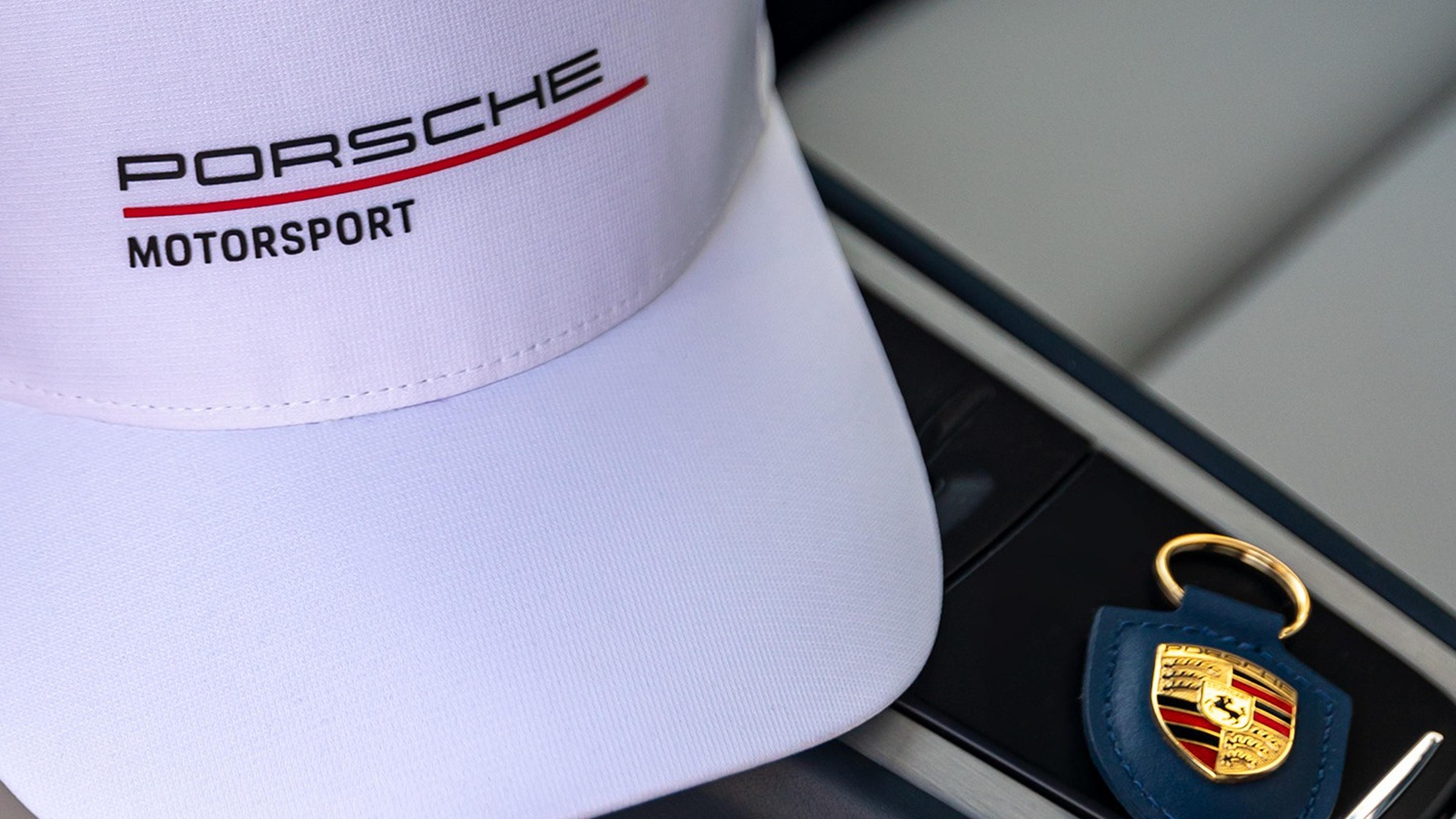 A white cap with "Porsche Motorsport" logo next to a Porsche-branded keychain with a golden ring on a car seat.