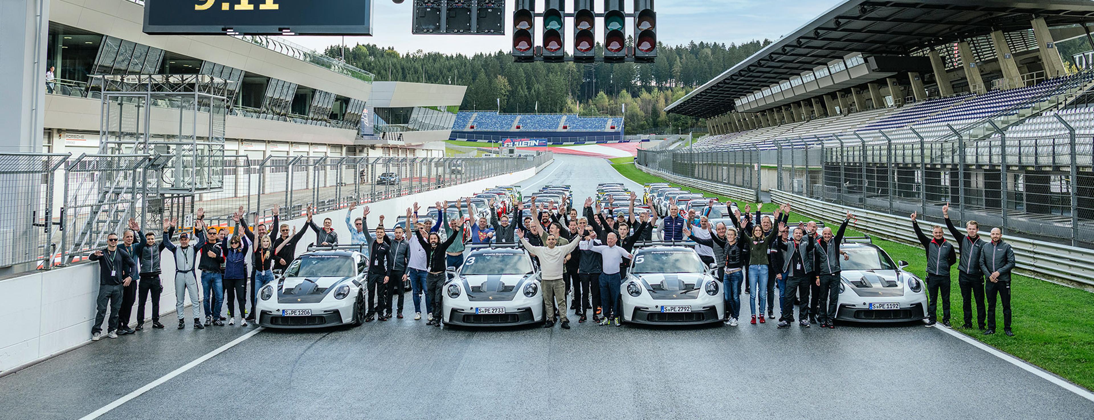 Eine Gruppe von Menschen steht neben drei weißen Sportwagen auf einer Rennstrecke und hebt die Arme; im Hintergrund sind Tribünen zu sehen.