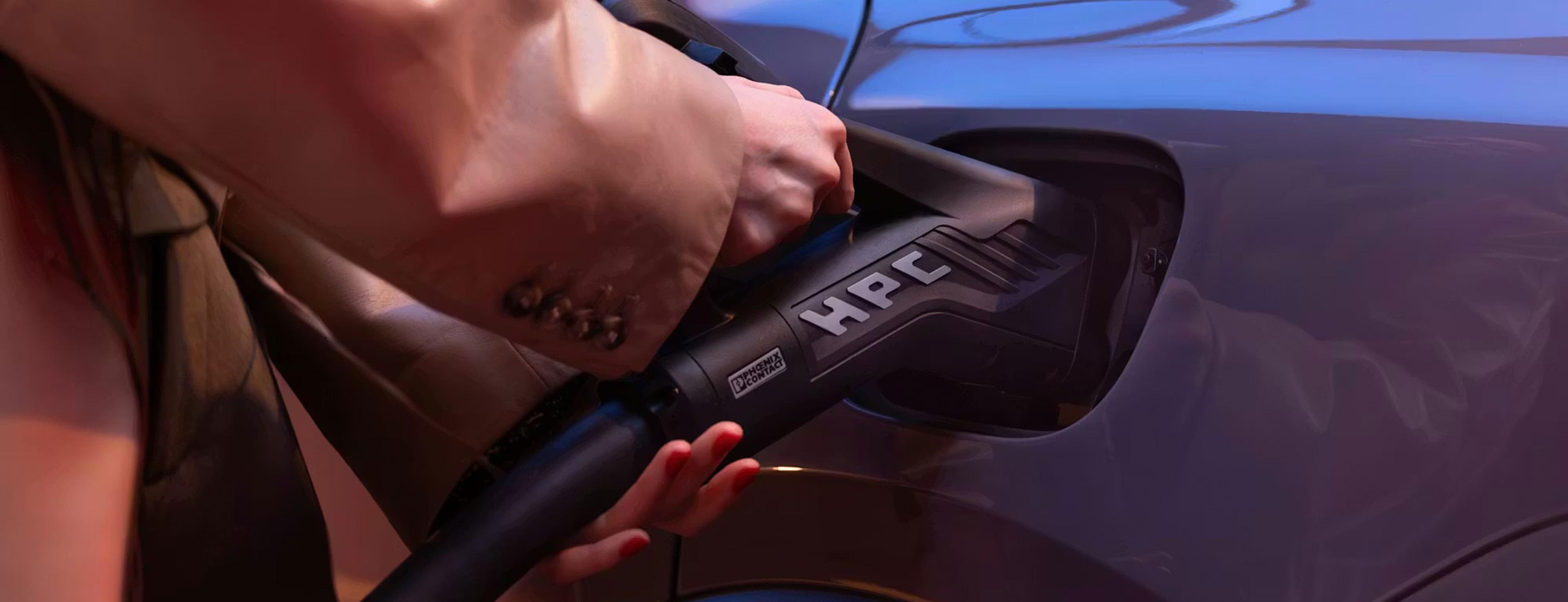 Eine Person in einem beigen Mantel steckt einen HPC-Ladestecker in die Ladebuchse eines blauen Autos.