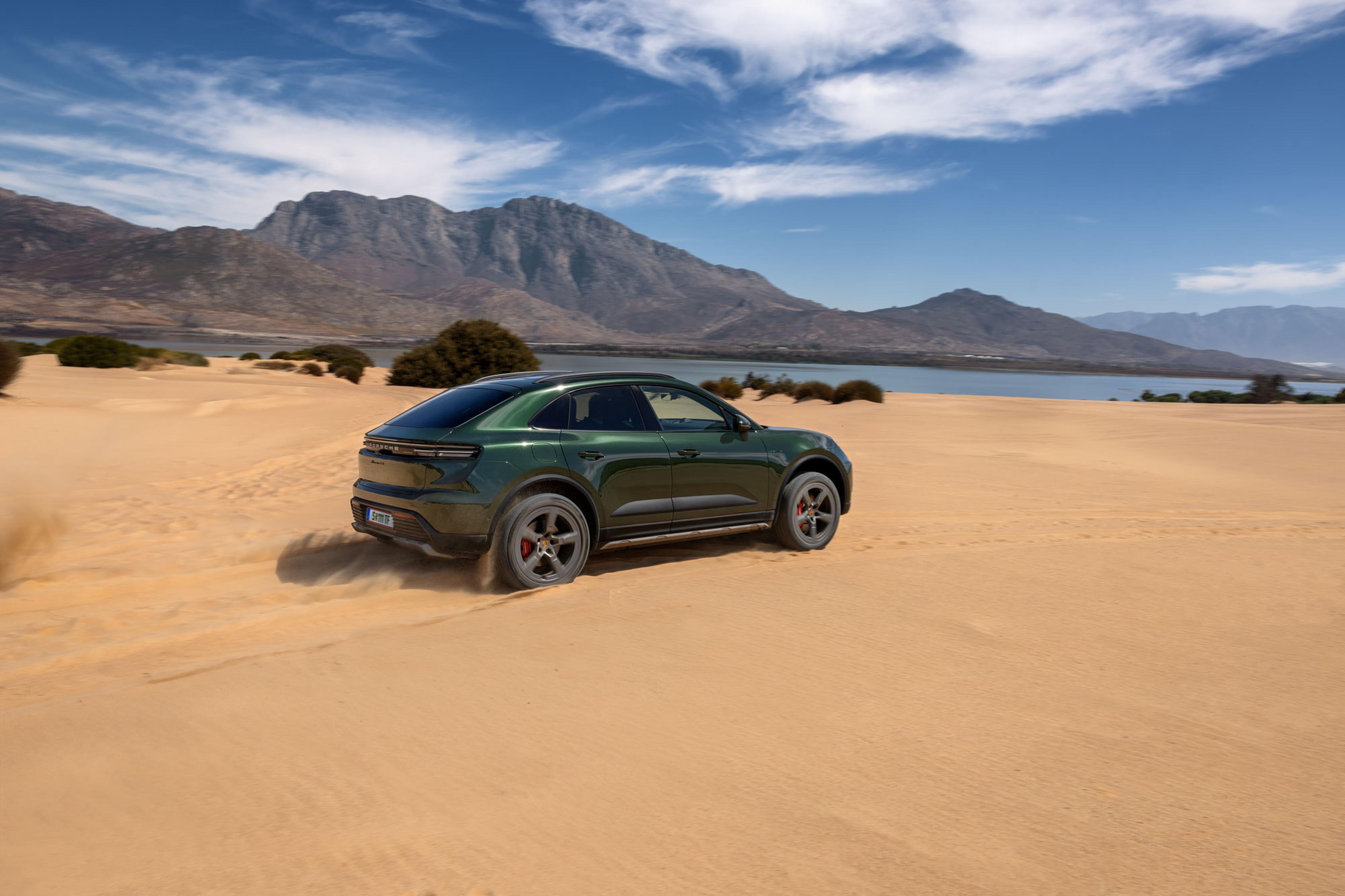 Ein grüner Porsche fährt durch sandiges Gelände, mit Bergen im Hintergrund unter einem teils bewölkten Himmel.