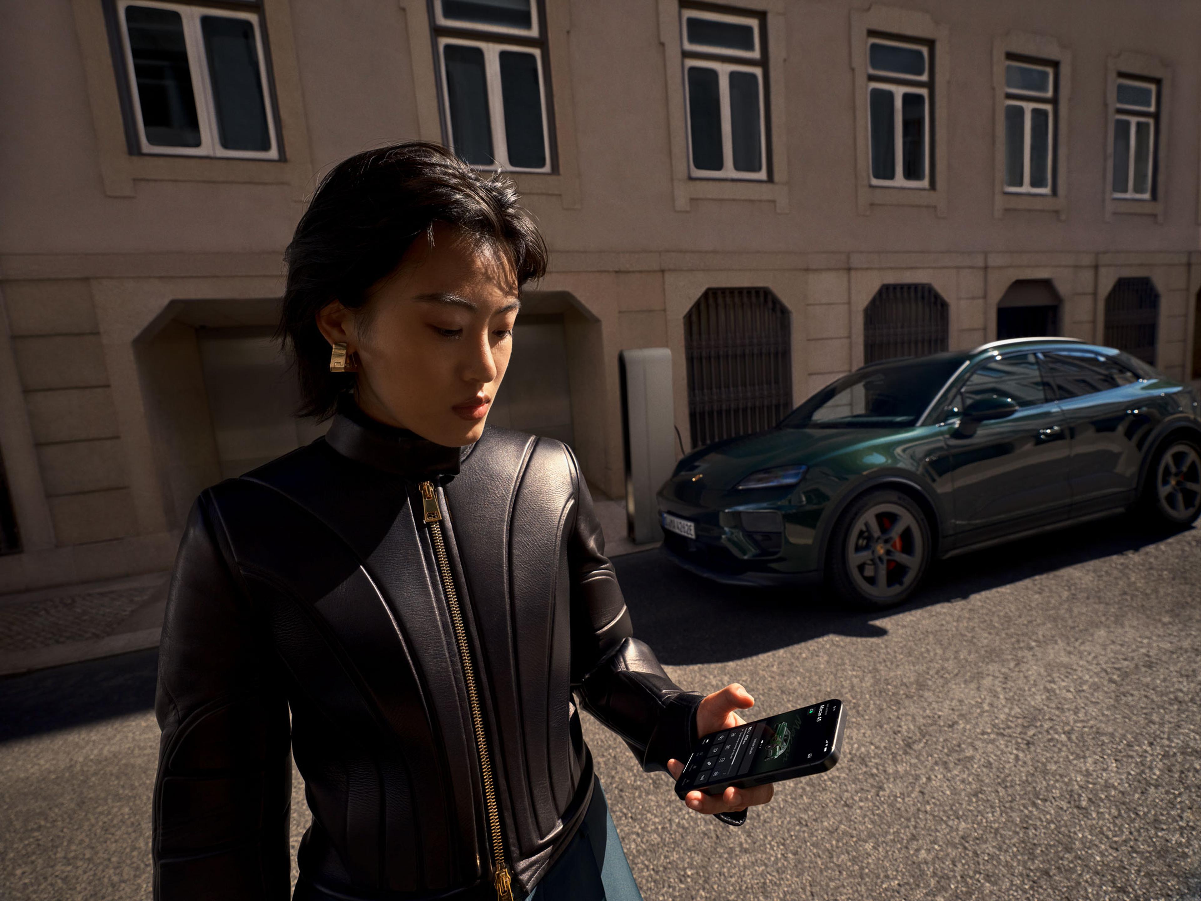 Eine Person in einer schwarzen Lederjacke betrachtet auf einer sonnigen Straße ein Smartphone, mit einem dunkelgrünen Auto und einem Gebäude im Hintergrund.