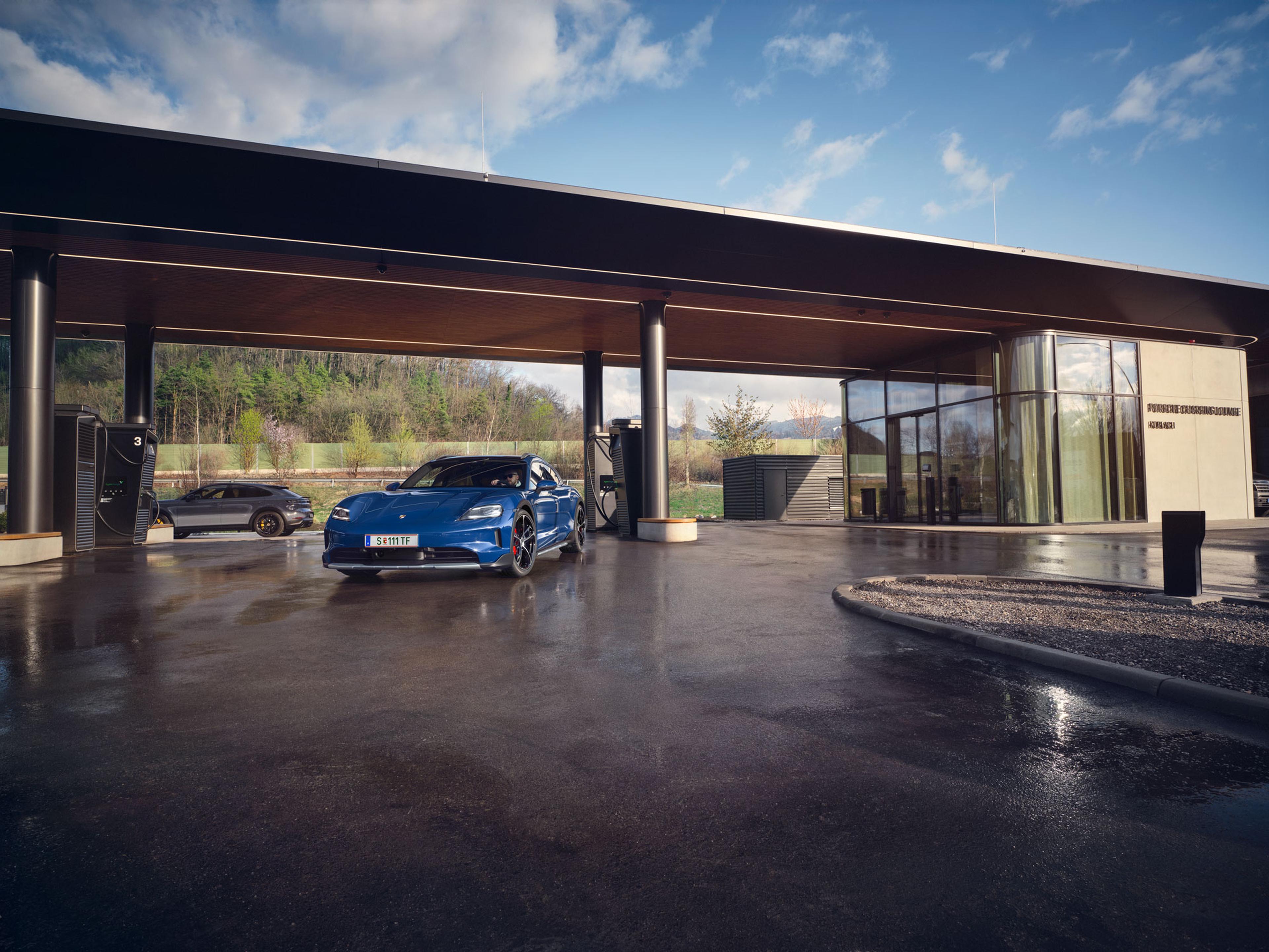 Ein blauer Porsche Taycan, geparkt an einer modernen Ladestation, mit einem eleganten Gebäude und einer grünen Landschaft im Hintergrund.