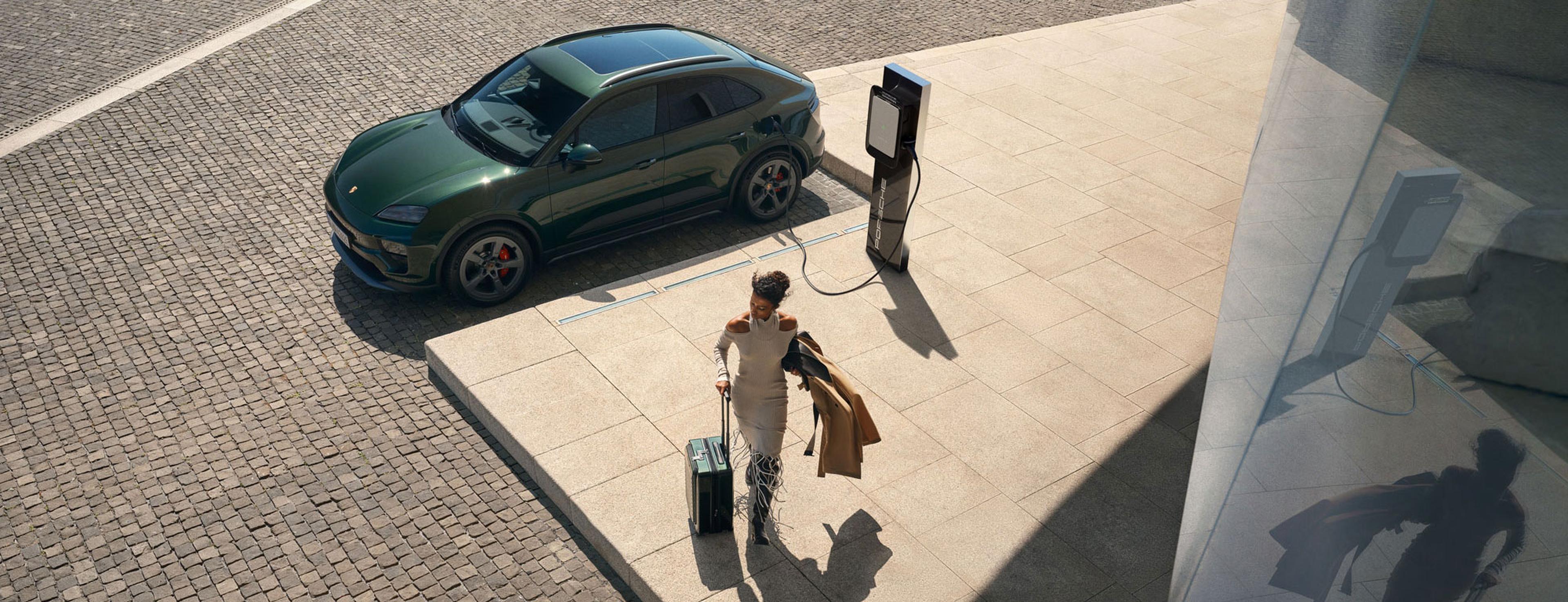 Ein violetter Sportwagen mit elegantem Design und modernen Scheinwerfern ist an einer Elektro-Ladestation geparkt; in der Nähe geht eine Person mit einem Koffer an einem Elektrofahrzeug vorbei, das gerade geladen wird und auf einer Kopfsteinpflasterstraße steht.