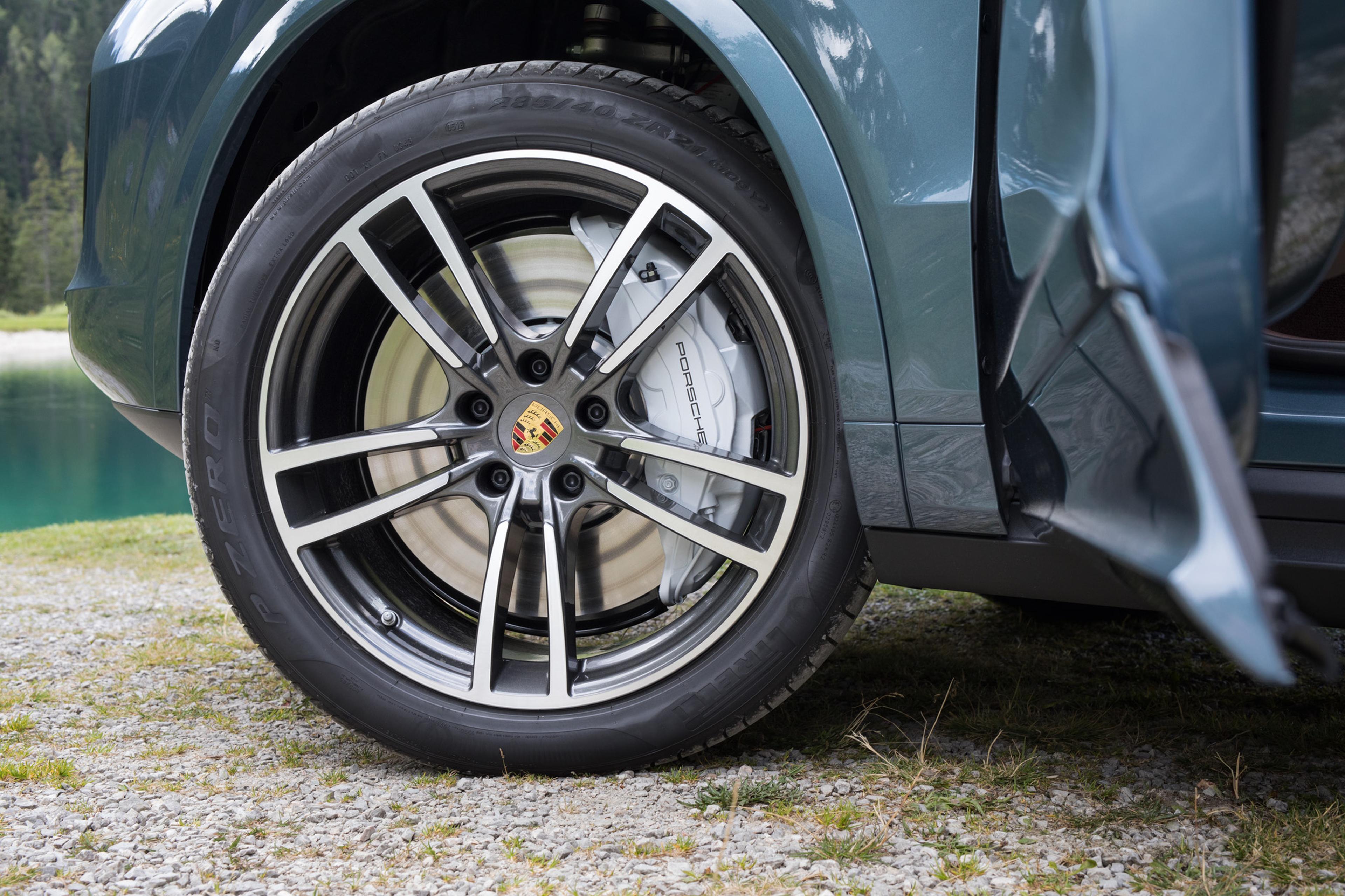 Hinterrad eines Autos mit Leichtmetallfelge und Porsche-Logo auf einem Kiesweg, mit Blick auf einen grünen See und einen bewaldeten Hintergrund.